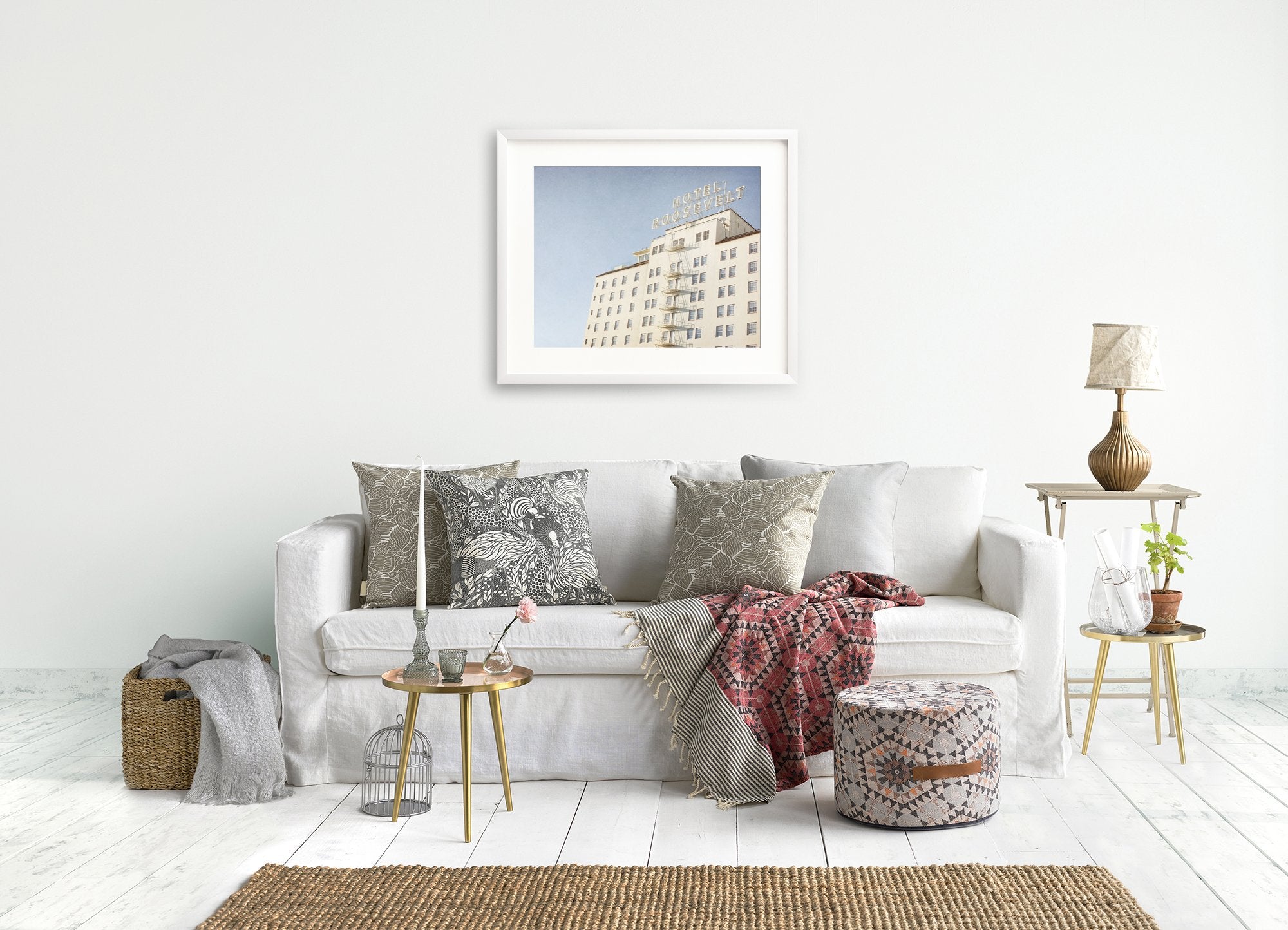 A cozy living room features a white sofa adorned with various patterned cushions and a red patterned throw blanket. Two side tables hold potted plants, a birdcage, and a lamp. A framed Retro Hollywood Print, 'Night in Tinseltown' by Offley Green hangs above the sofa. A woven rug lies on the white wooden floor.