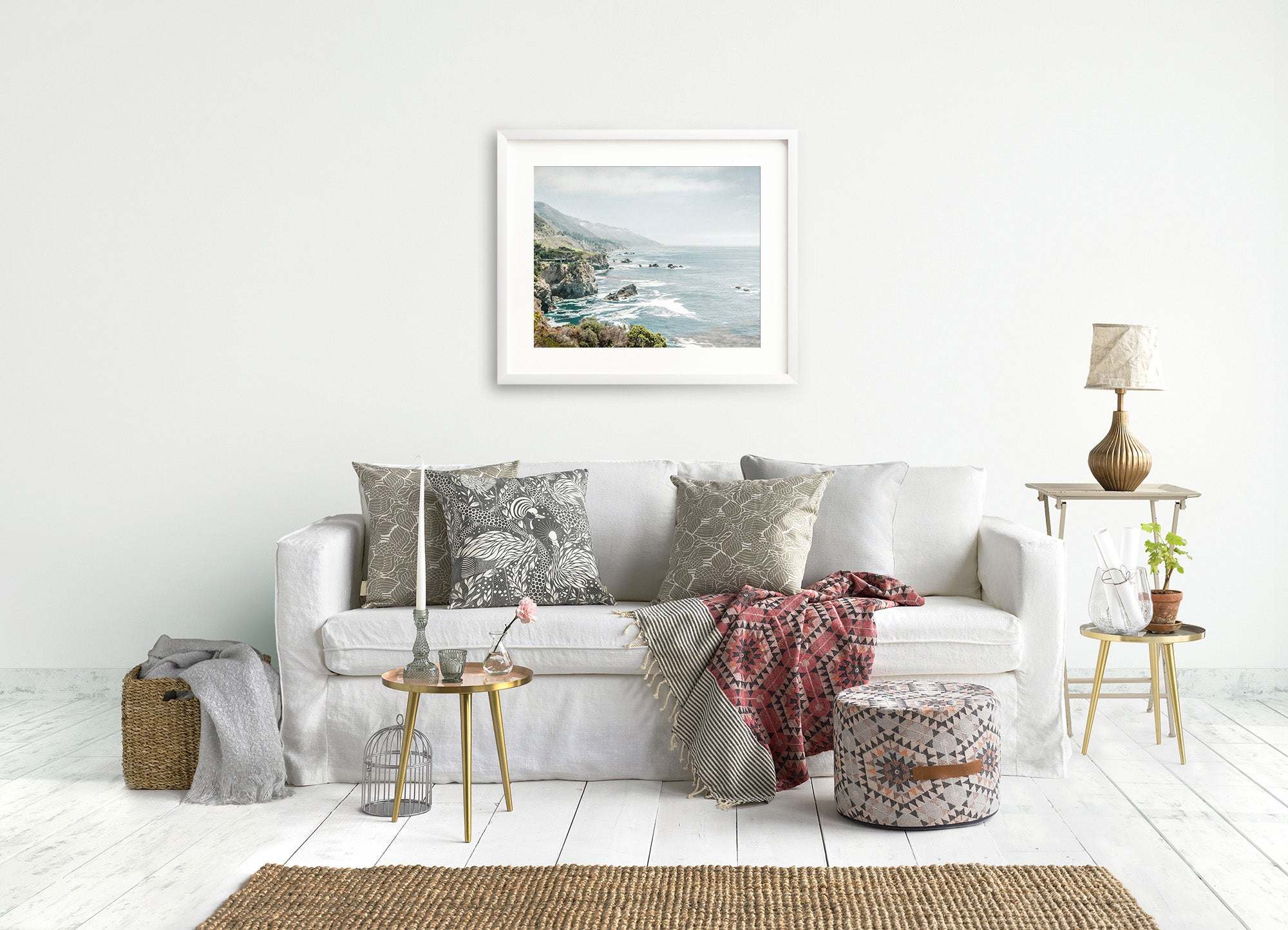 A cozy living room with a white sofa decorated with patterned cushions, a side table with a lamp, a basket, and an Offley Green Big Sur Landscape Print of 'Rocky Rocks' on the wall.