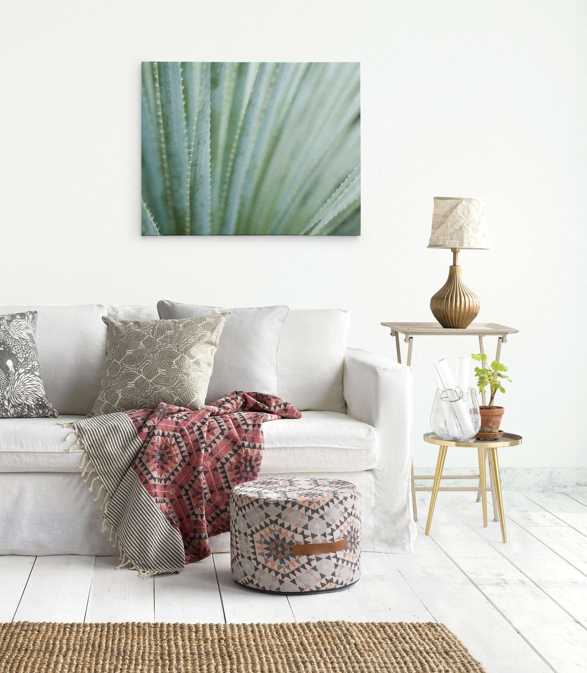 A cozy living room corner with a white sofa adorned with various patterned pillows and a red throw, a small wooden side table with a lamp, and an Offley Green 'Strands and Spikes' Abstract Green Botanical Canvas Wall Art on the wall.