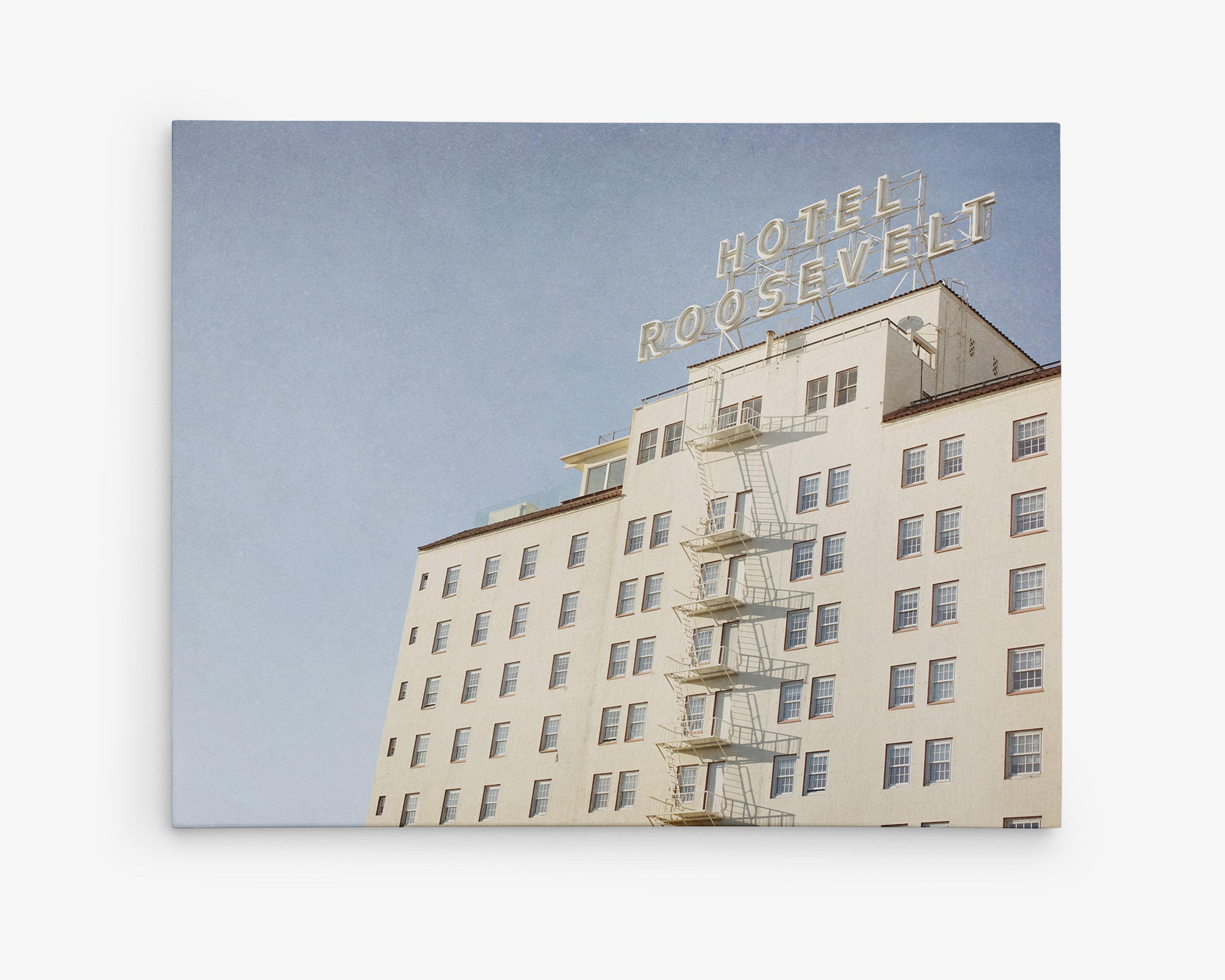A photograph of the Offley Green Retro Hollywood Canvas Wall Art, 'Night in Tinseltown' under a clear sky, showing its white facade and a prominent neon sign on top, with an exterior fire escape on the side.