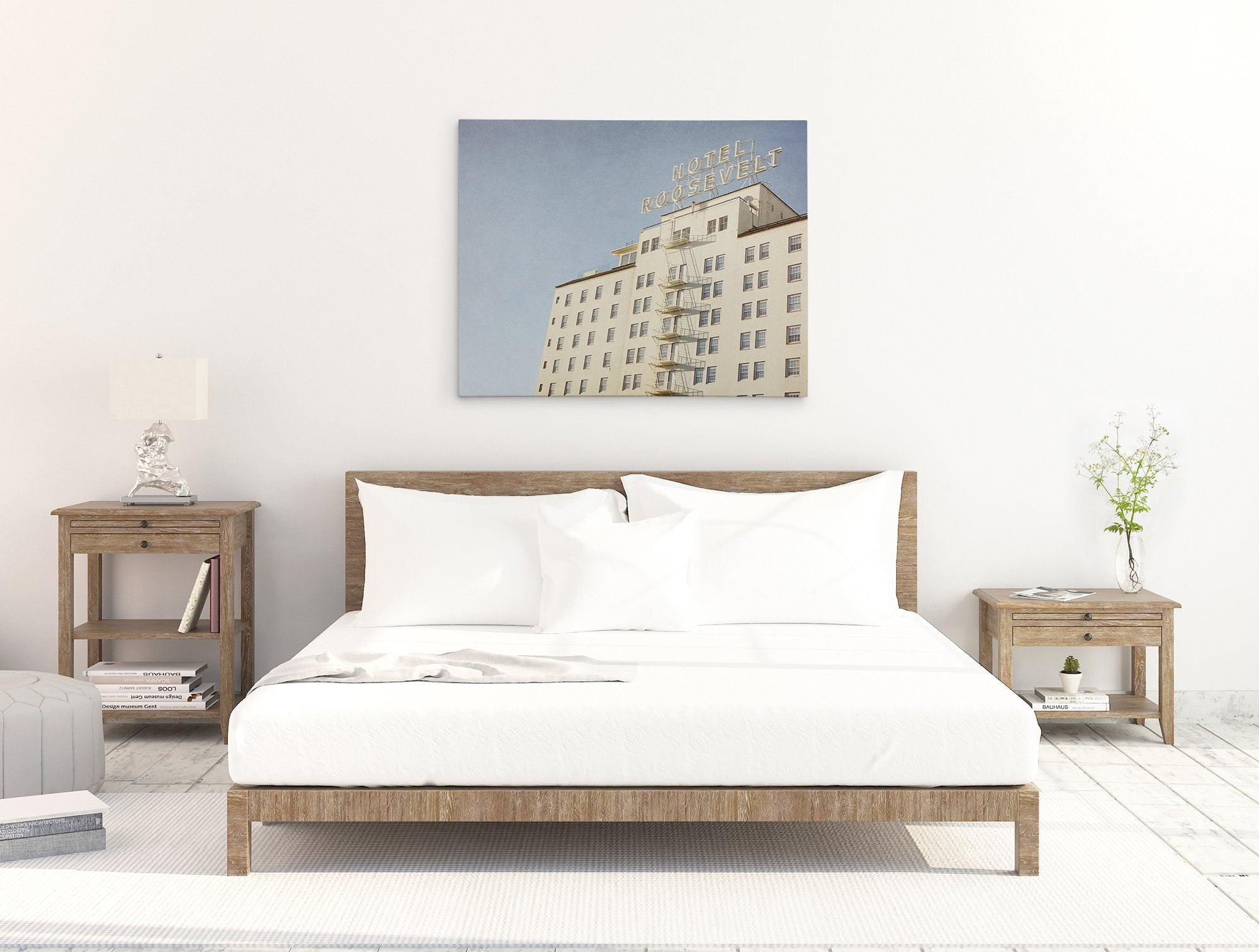 A minimalist bedroom featuring a large bed with white bedding centered between two wooden nightstands, under a Offley Green &#39;Night in Tinseltown&#39; Retro Hollywood Canvas Wall Art of the Roosevelt Hotel Hollywood on a white wall.