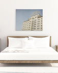 A minimalist bedroom featuring a large bed with white bedding centered between two wooden nightstands, under a Offley Green 'Night in Tinseltown' Retro Hollywood Canvas Wall Art of the Roosevelt Hotel Hollywood on a white wall.