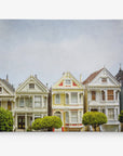 A row of five colorful Victorian houses, known as the Painted Ladies, stands closely together in San Francisco. Each house has distinct ornate details and pastel colors ranging from yellow and blue to red and beige. Two trimmed bushes are in front of the houses under a cloudy sky, like Offley Green's San Francisco Canvas Wall Art, 'Painted Ladies' come to life.