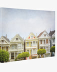 The San Francisco Canvas Wall Art, 'Painted Ladies' by Offley Green depicts a row of colorful Victorian houses, known as the Painted Ladies in San Francisco, with intricate architectural details. Each house features ornate trim, bay windows, and gabled roofs. In the foreground, neatly trimmed trees add greenery to the scene. The sky is clear and light blue.