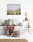 A cozy living room with a white sofa adorned with patterned throw pillows and a colorful blanket. A small table on the right holds a lamp and a plant. Above the sofa, an Offley Green San Francisco Canvas Wall Art, 'Painted Ladies', adds charm. A woven rug and a patterned pouf complete the space.