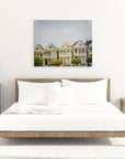 A minimalistic bedroom with a wooden bed frame and white bedding. Two matching wooden nightstands with lamps and decorative items flank the bed. A San Francisco Canvas Wall Art, 'Painted Ladies' by Offley Green hangs on the white wall above the bed. The floor is light-colored wood.