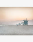 A Pink Coastal Print, 'Lifeguard Tower' by Offley Green stands isolated on a sandy beach in Venice with a calm sea in the background under a soft gradient sky of pastel colors at dawn or dusk.