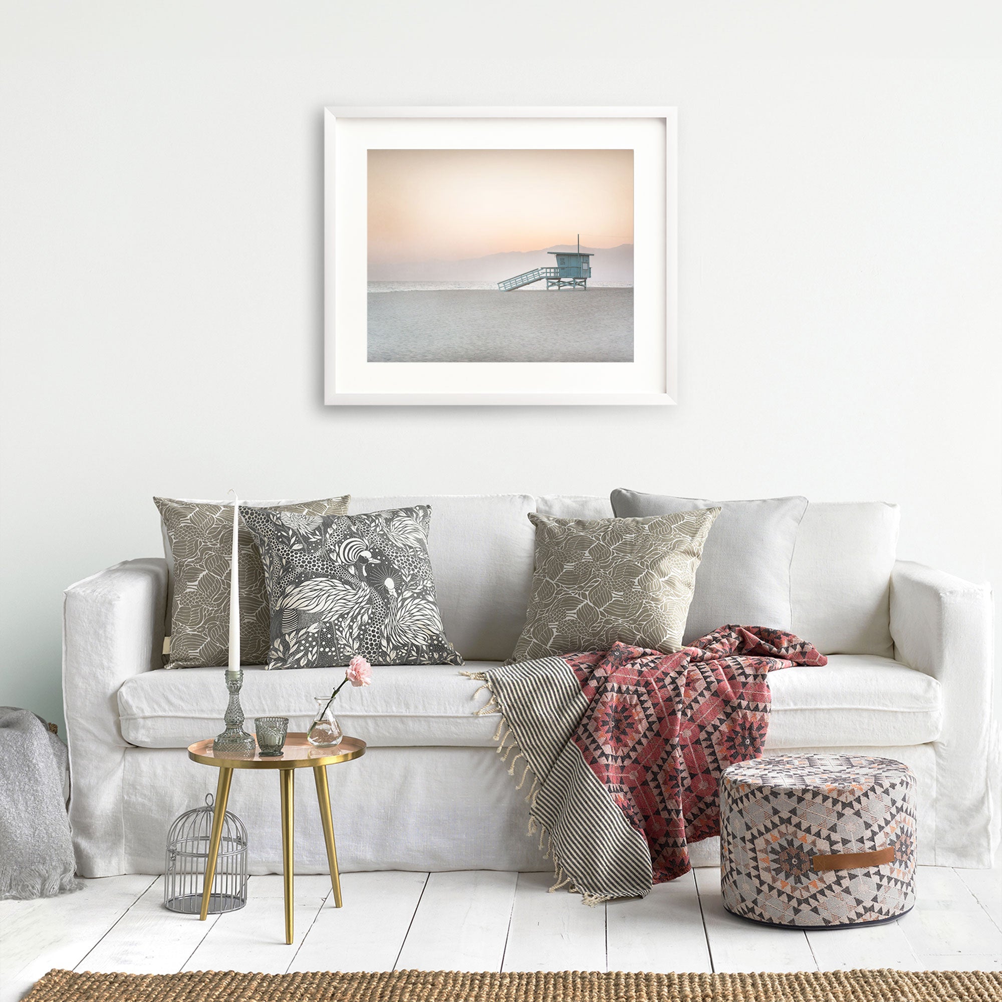 A cozy living room scene with a white sofa adorned with decorative pillows, a red patterned throw blanket, and a small table with vases and books. A tranquil Pink Coastal Print, &#39;Lifeguard Tower&#39; photograph of Venice hangs on the wall from Offley Green.