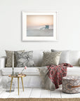 A cozy living room scene with a white sofa adorned with decorative pillows, a red patterned throw blanket, and a small table with vases and books. A tranquil Pink Coastal Print, 'Lifeguard Tower' photograph of Venice hangs on the wall from Offley Green.
