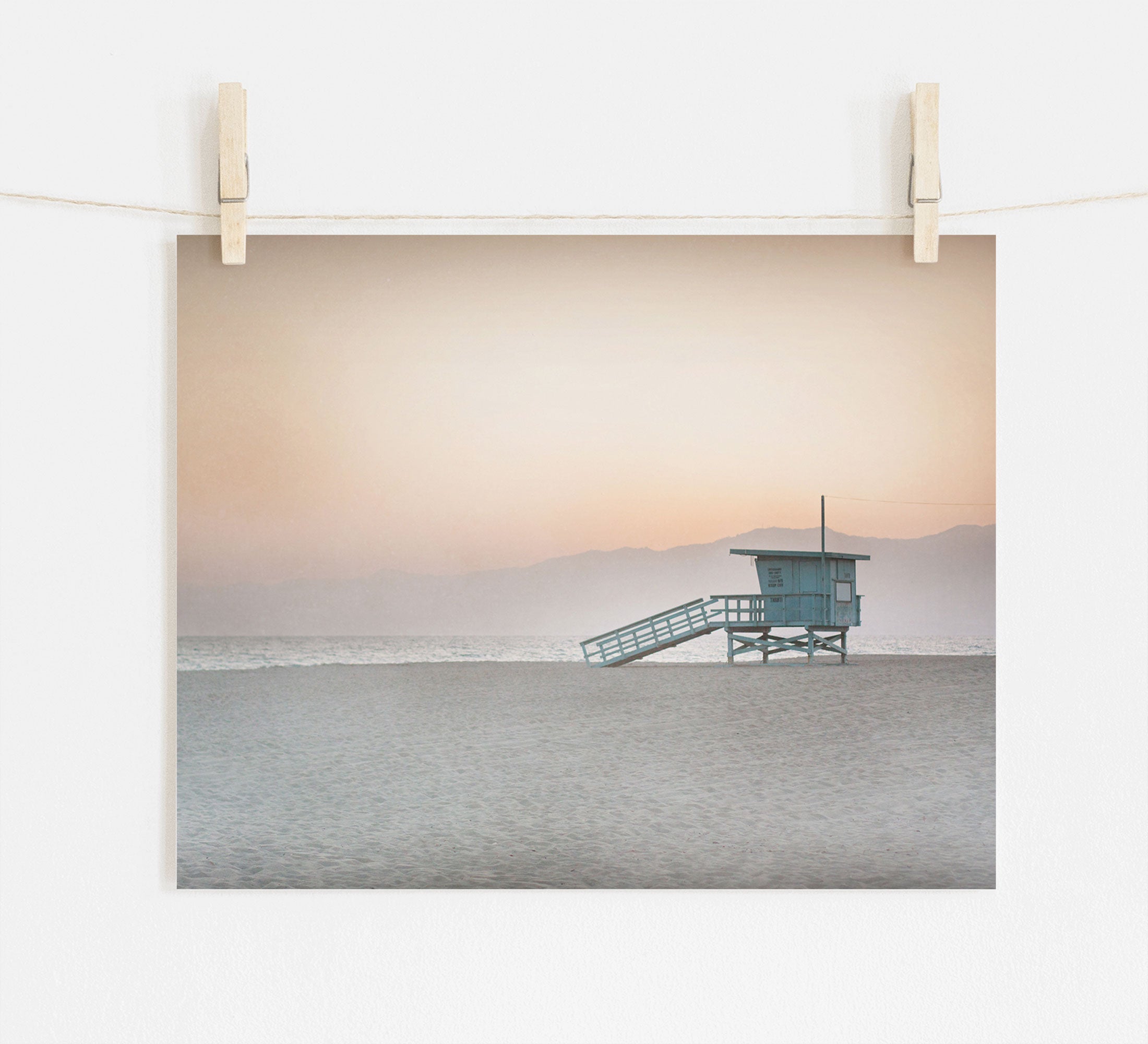 A serene beach scene at dusk printed on archival photographic paper, hung by clips on a string against a white wall. It features the Offley Green Pink Coastal Print, &#39;Lifeguard Tower&#39; on an empty sandy beach near Venice and Santa Monica.