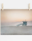 A serene beach scene at dusk printed on archival photographic paper, hung by clips on a string against a white wall. It features the Offley Green Pink Coastal Print, 'Lifeguard Tower' on an empty sandy beach near Venice and Santa Monica.