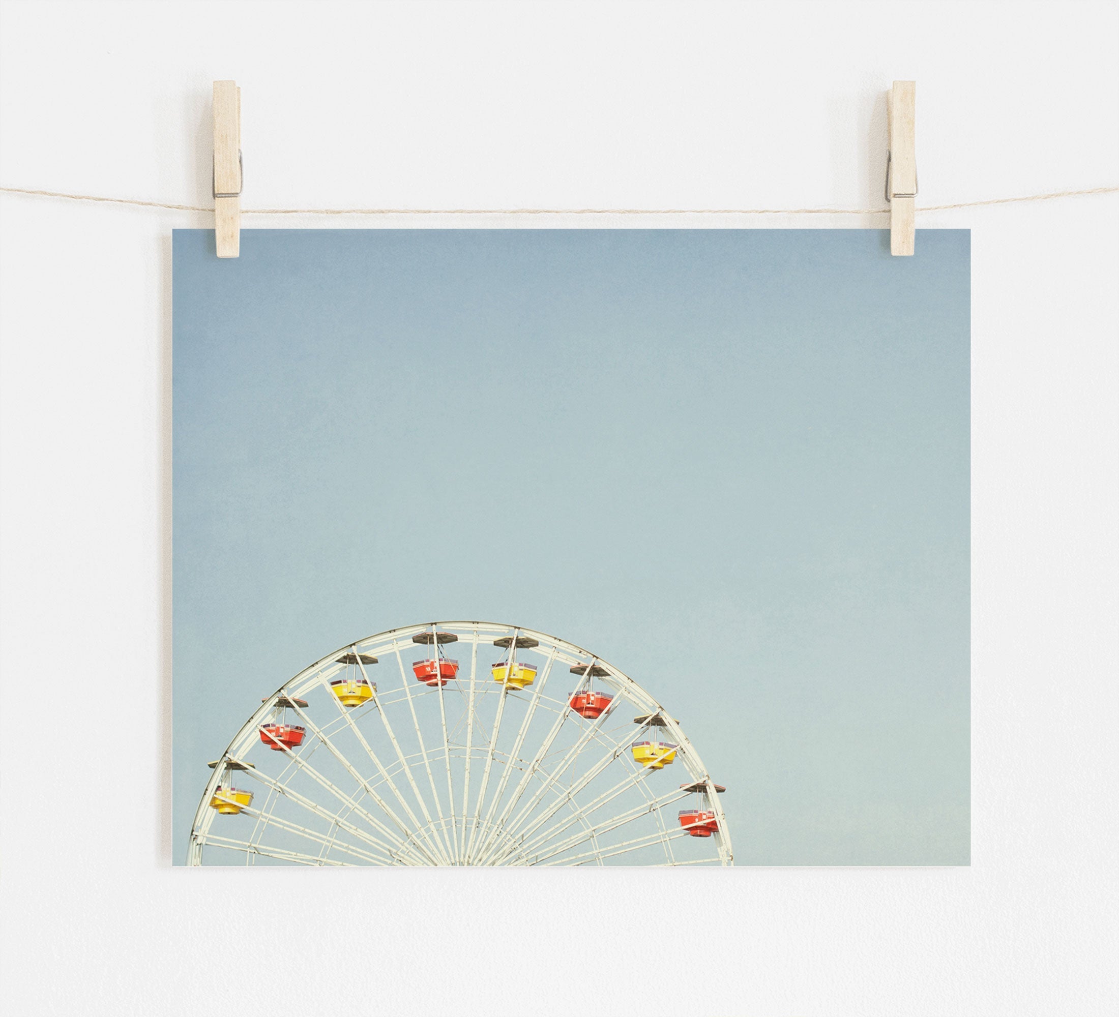 A photograph pinned by wooden clothespins on a string, depicting the top half of a ferris wheel with colorful cabins at Santa Monica Pier, set against a plain light blue background - Blue Minimalist Wall Decor, 'Ferris Blue' by Offley Green.