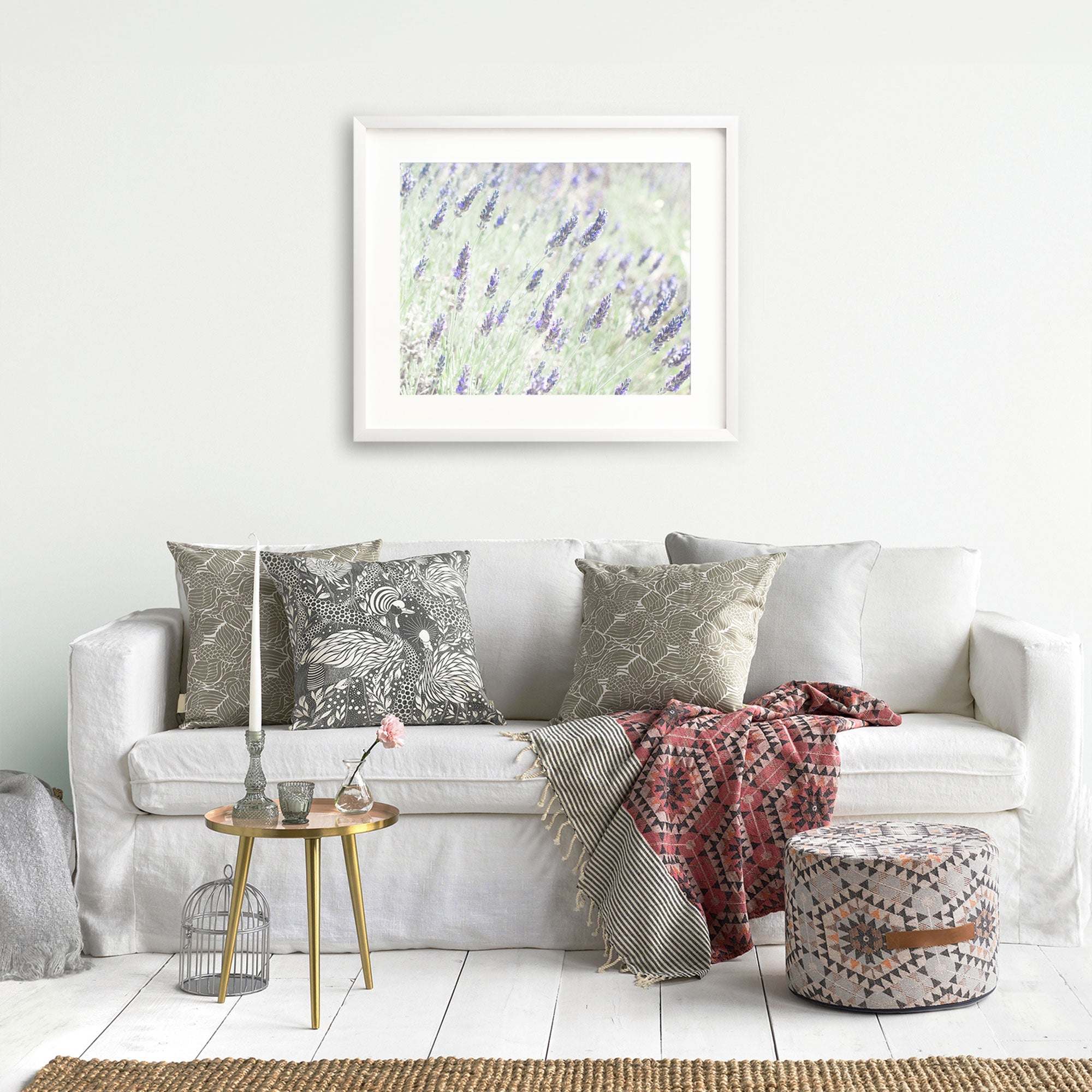 A cozy living room with a white sofa adorned with patterned pillows, a red patterned throw blanket, a small round gold table with decorative items, and Offley Green&#39;s &#39;Lavender for LaLa&#39; Floral Purple Print above the sofa.