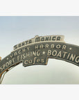 The image shows the iconic Santa Monica Pier arch sign with the text "yacht harbor, sport fishing, boating, cafes" against a clear sky on the Los Angeles California Print, 'Santa Monica Pier Blues' by Offley Green.