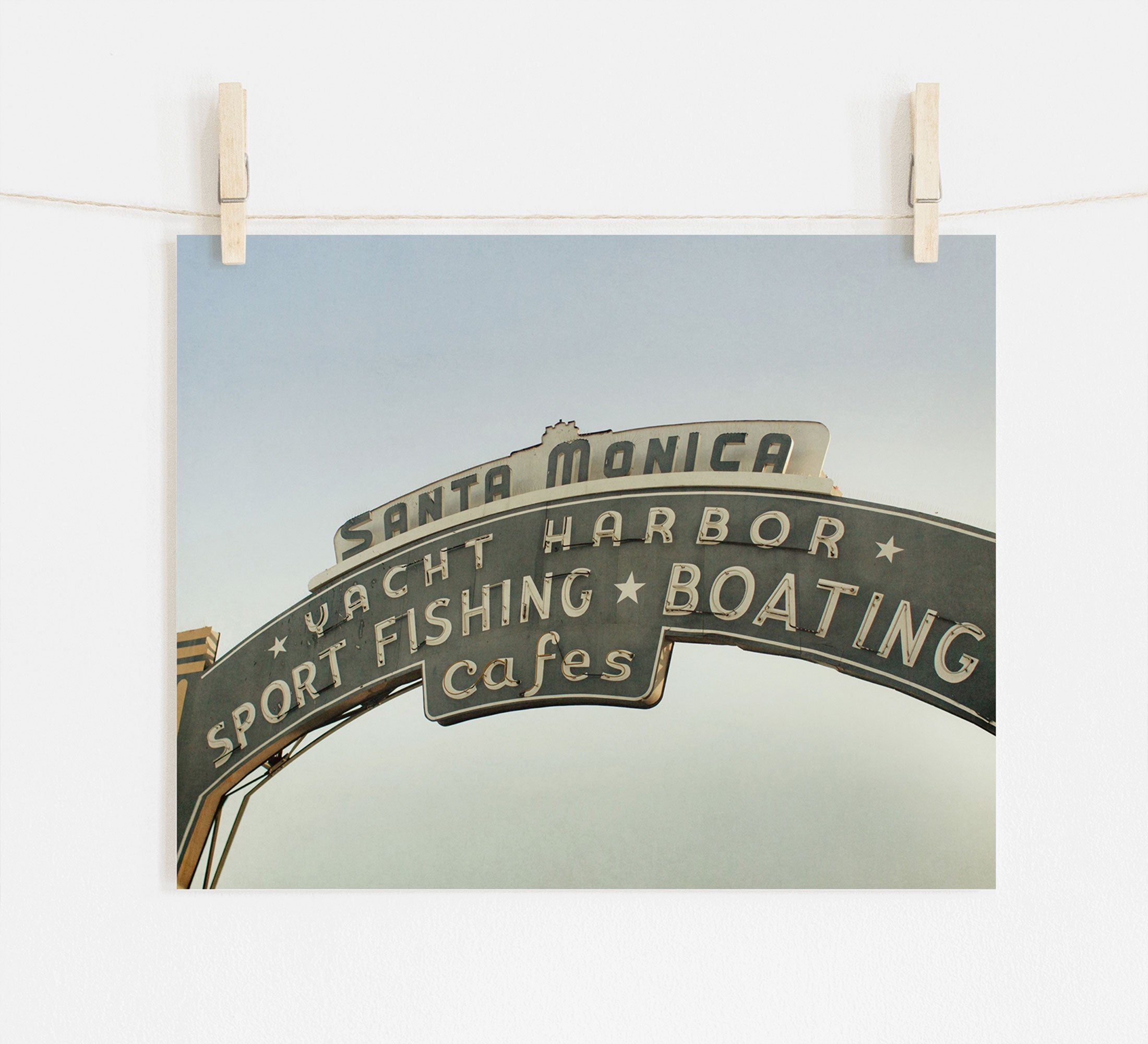 A photograph of the Los Angeles California Print, 'Santa Monica Pier Blues' by Offley Green, with words "sport fishing boating cafes", pinned to a white wall with wooden clothespins, now showcased as beach town wall art.