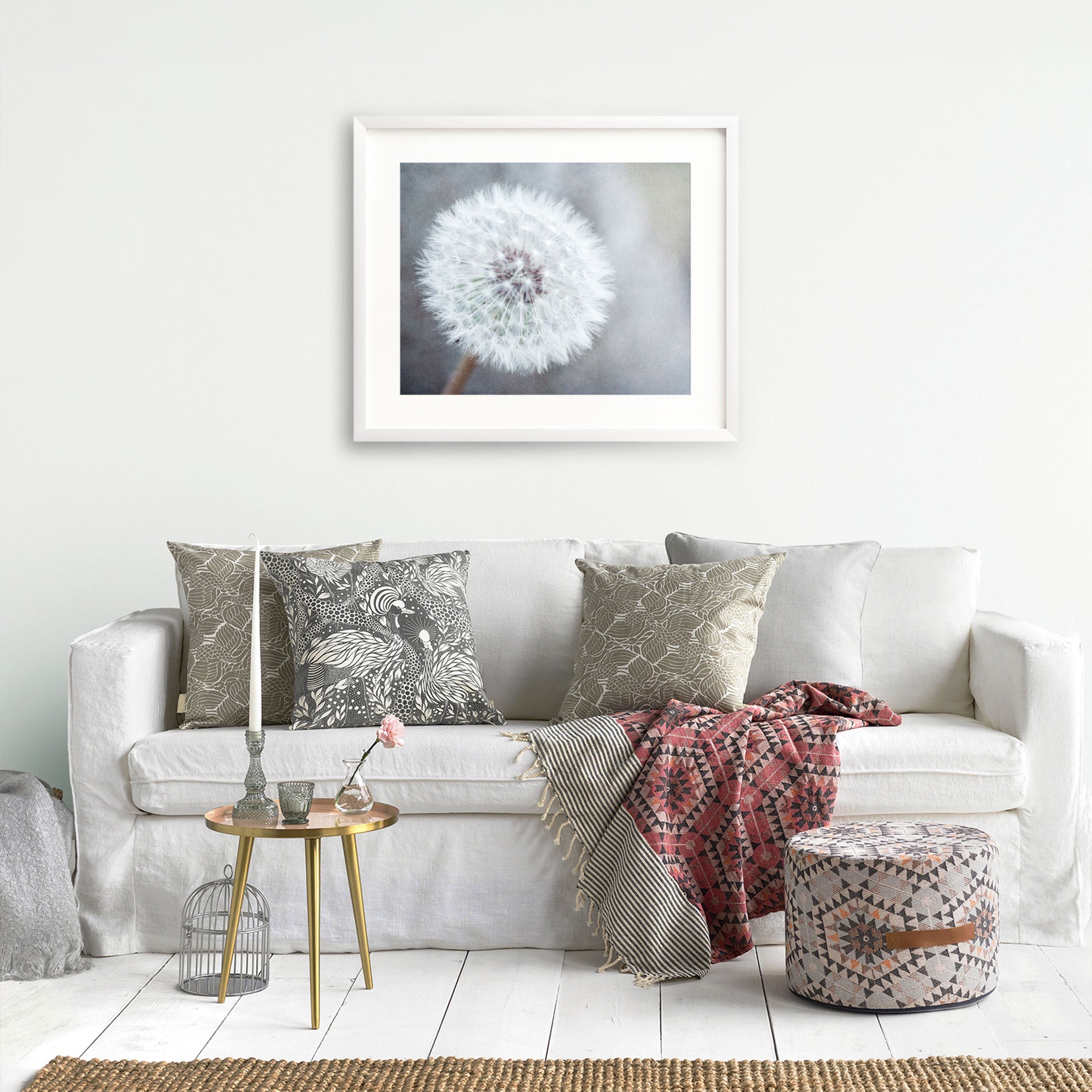 A cozy living room scene featuring a white sofa with decorative pillows, an unframed Neutral Grey Floral Print of a dandelion on the wall, a small round table with books, a birdcage, and 'Dandelion King' by Offley Green.