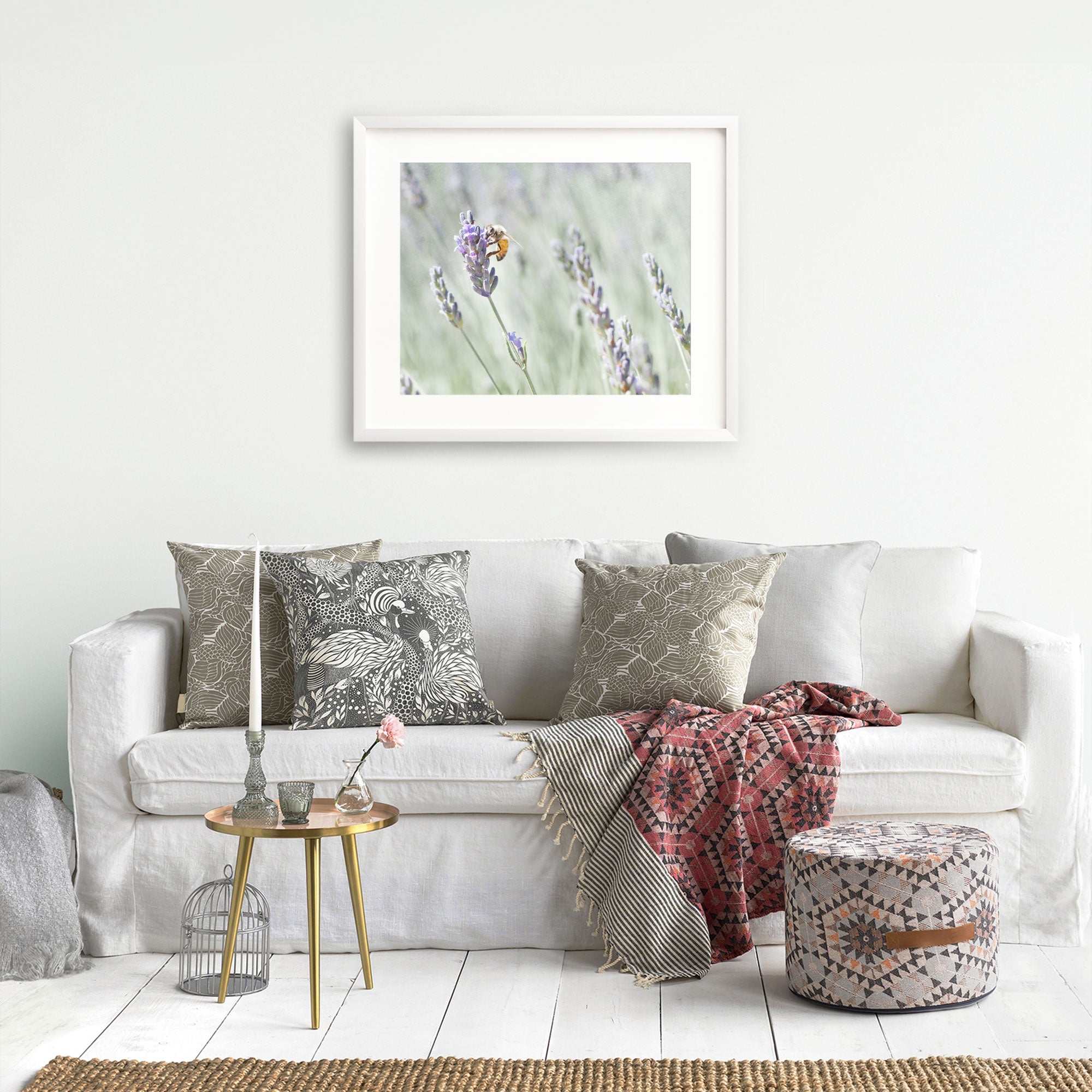 A cozy living room featuring a white sofa adorned with stylish patterned cushions, a red throw blanket, and a small round table. A framed Offley Green Rustic Floral Print, &#39;Lavender for Bees&#39;, printed on archival photographic paper.