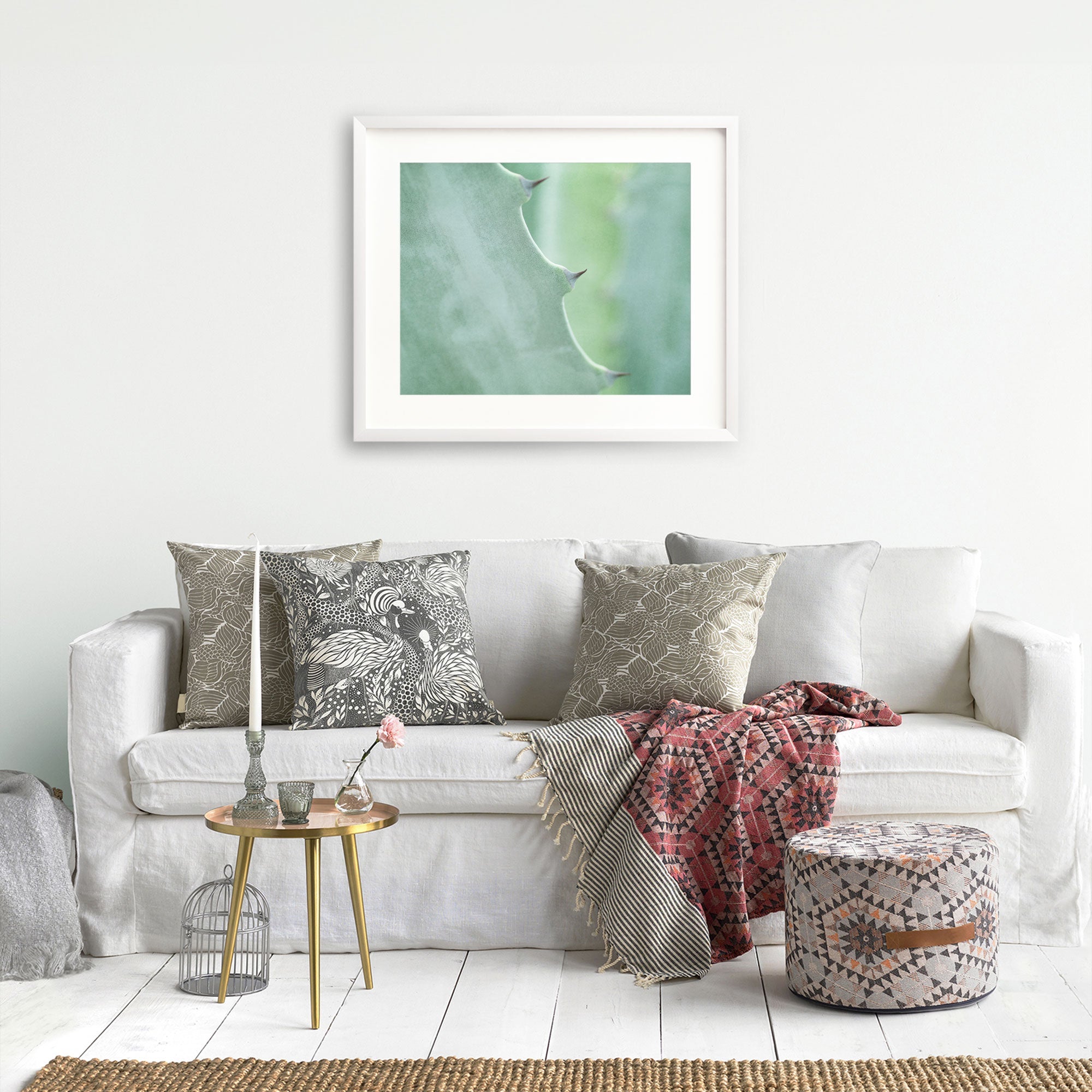 A cozy living room scene with a white couch adorned with patterned pillows, a red throw blanket, a small round table with a book and an Aloe Vera plant, a framed abstract art piece featuring the Mint Green Botanical Print 'Aloe Vera Spikes' from Offley Green.