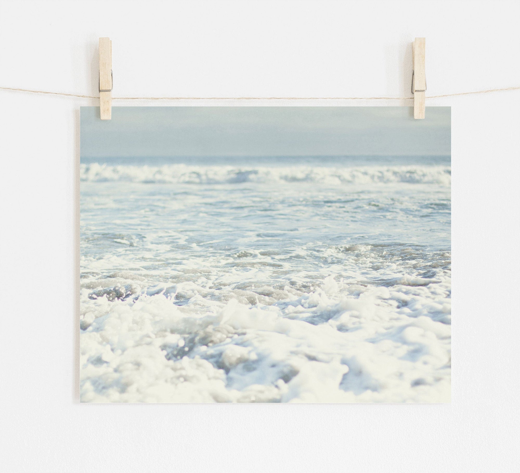 A photograph of a serene ocean scene at Pfeiffer Beach is displayed, clipped to a string with wooden clothespins. The image captures gentle waves breaking on the shore, with soft light reflecting on the frothy water under a clear sky, all printed on archival photographic paper. This is the Ocean Waves Print, 'Fresh Surf' by Offley Green.