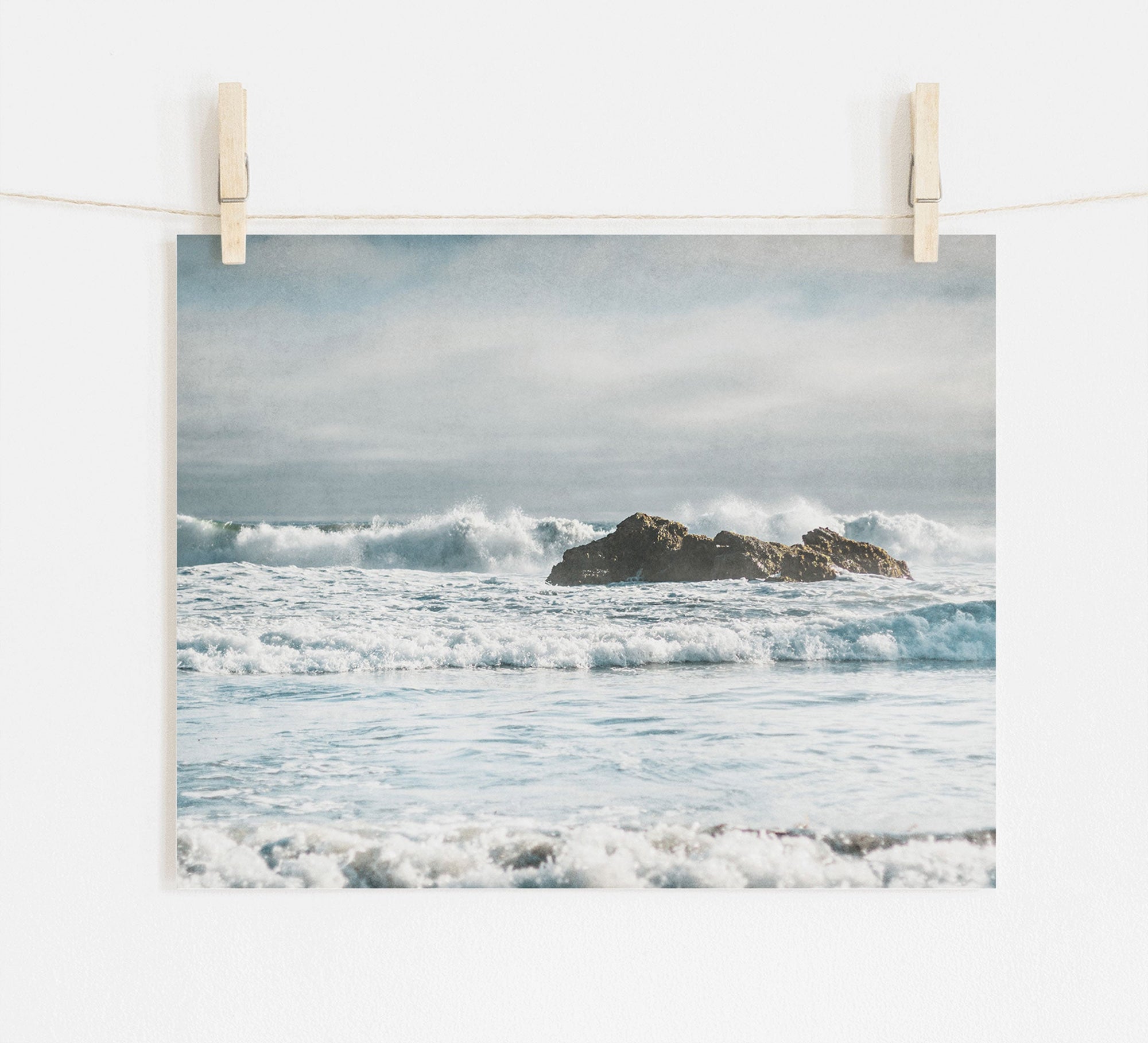 A photograph of a moody seascape with waves crashing against a small, rocky island in the center. The picture, Seascape Coastal Art, 'Surf and Rocks' by Offley Green, printed on archival photographic paper, is clipped to a string with two wooden clothespins, hanging against a white wall. The sky above is cloudy, creating a serene and tranquil atmosphere.