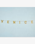 A minimalist representation with the Venice Beach Sign Print, 'Blue Venice' spelled out in yellow letters on small flags strung across a plain light blue background, printed on archival photographic paper by Offley Green.