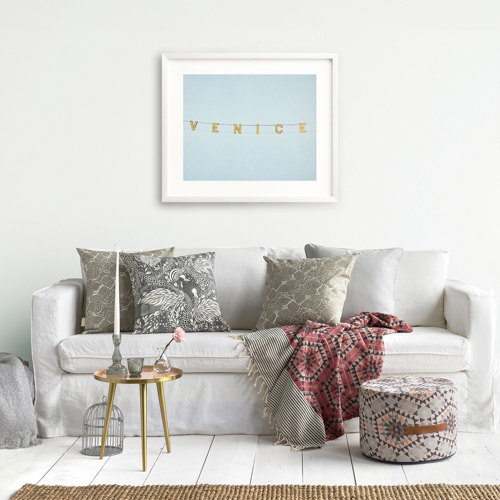 A cozy living room corner with a white sofa adorned with patterned pillows, a red blanket, and unframed California coastal photography prints on the wall above. A small side table with books and a Offley Green's Venice Beach Sign Print, 'Blue Venice'.