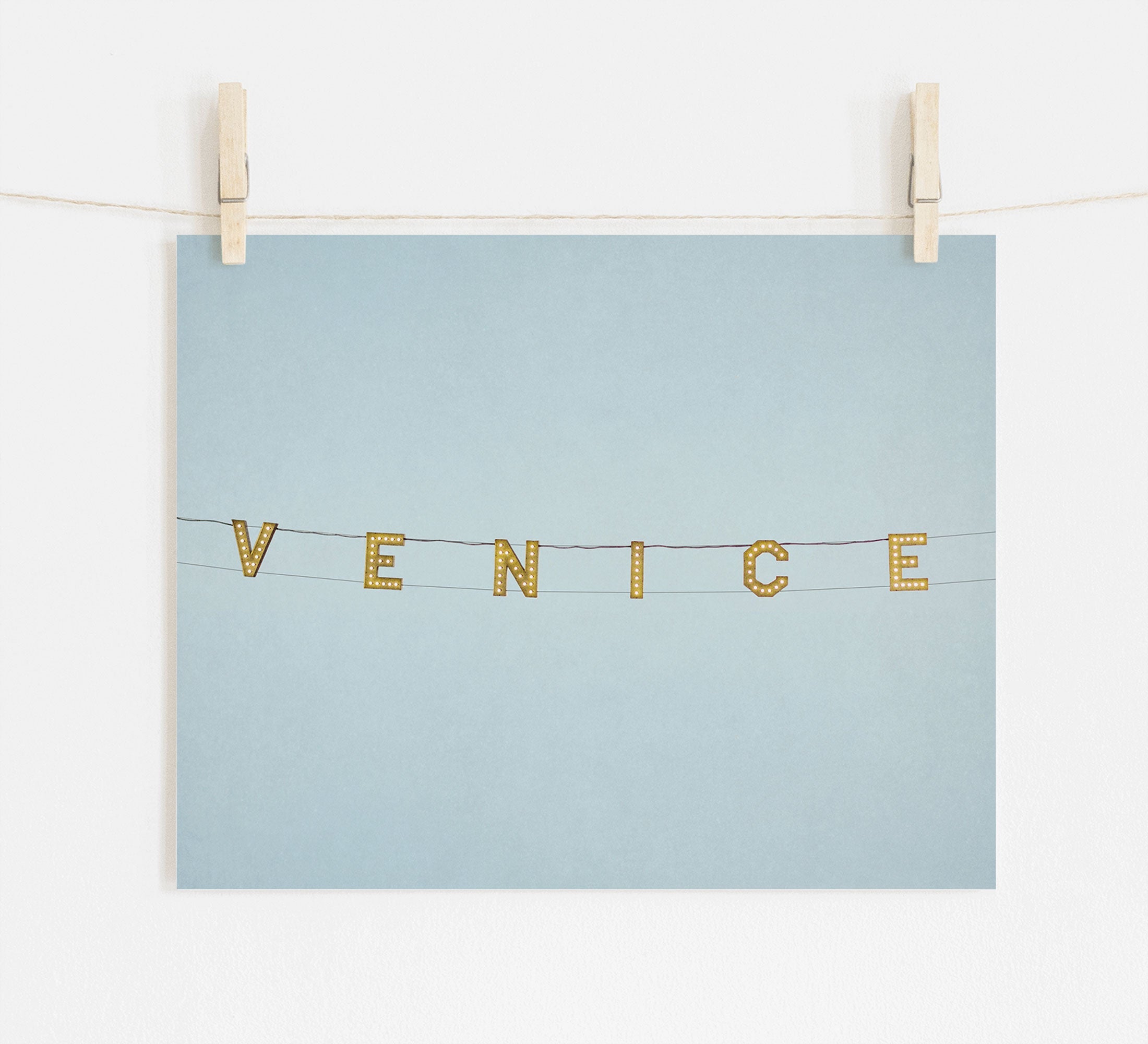 A light blue postcard, printed on archival photographic paper, is hung on a white wall by two wooden clips on a string, featuring the word &quot;Venice Beach Sign Print, &#39;Blue Venice&#39;&quot; in decorative gold letters strung horizontally from Offley Green.