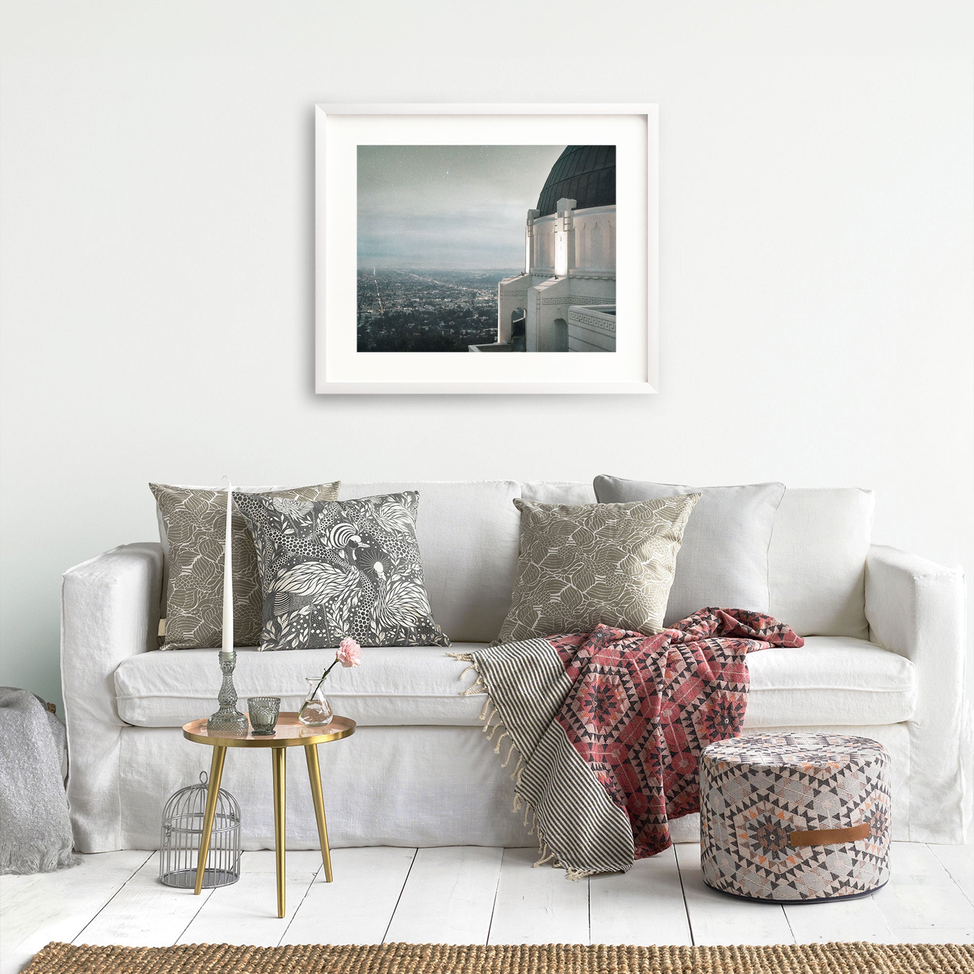 A minimalist living room featuring a white sofa adorned with patterned pillows, a small metallic side table with decor, a woven pouf, and an Offley Green archival photograph print of Griffith Observatory ('The Sky At Night') hanging on the wall.