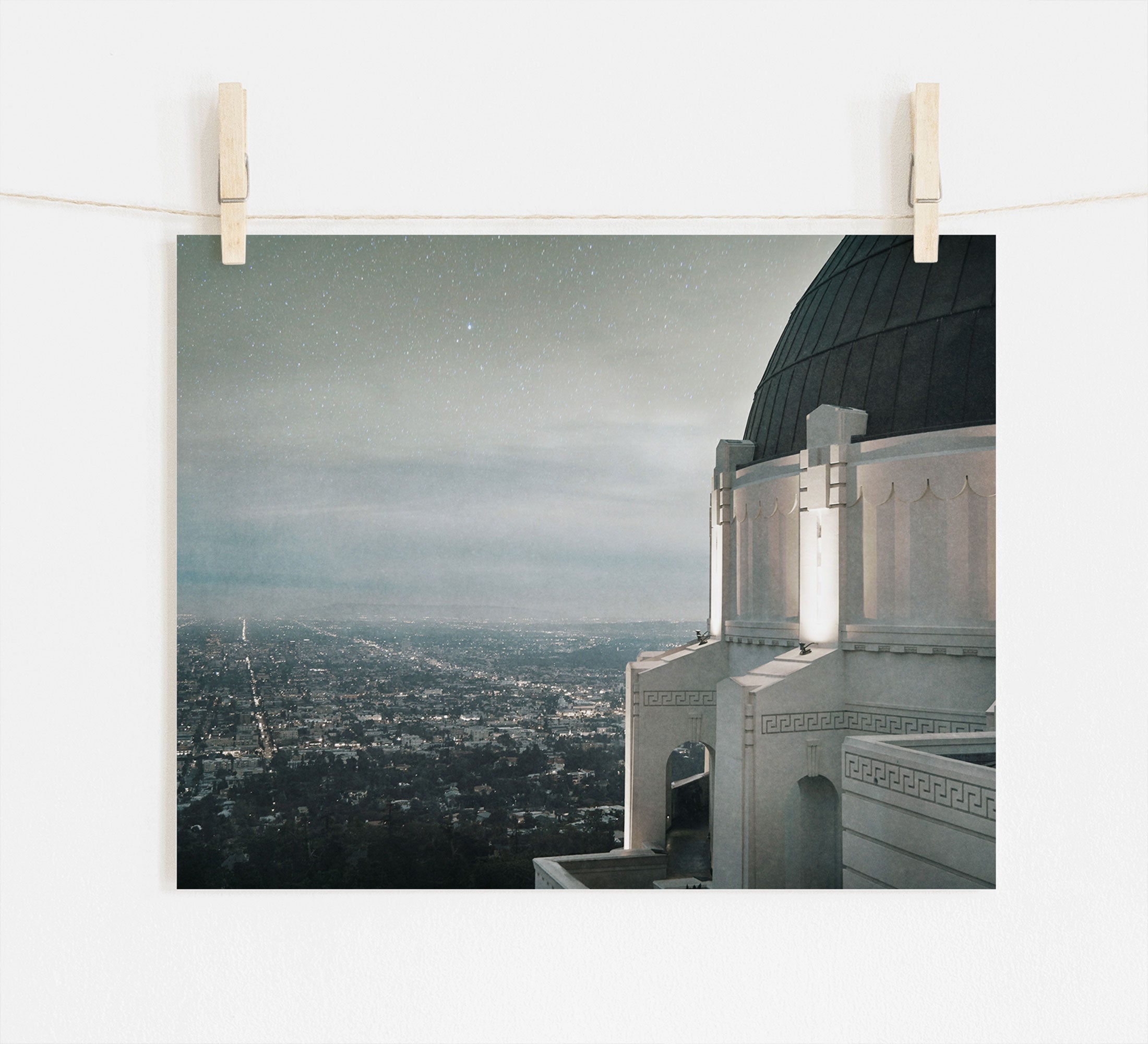 Photo of a cityscape at dusk pinned to a line, with Offley Green's Griffith Observatory Print, 'The Sky At Night' in the foreground and twinkling city lights below a starry sky.
