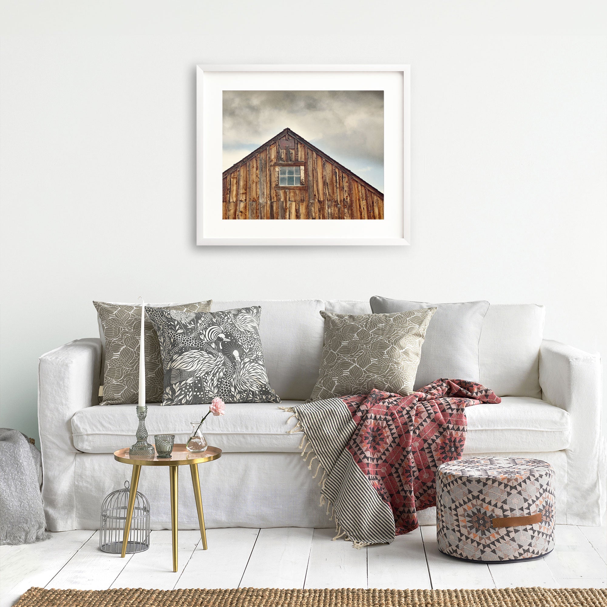 A cozy living room featuring a white sofa adorned with patterned throw pillows, a red and grey blanket, and a small round table with decorative items. Above the sofa, a framed photo of the Offley Green Farmhouse Rustic Print, 'Old Barn at Bodie'.