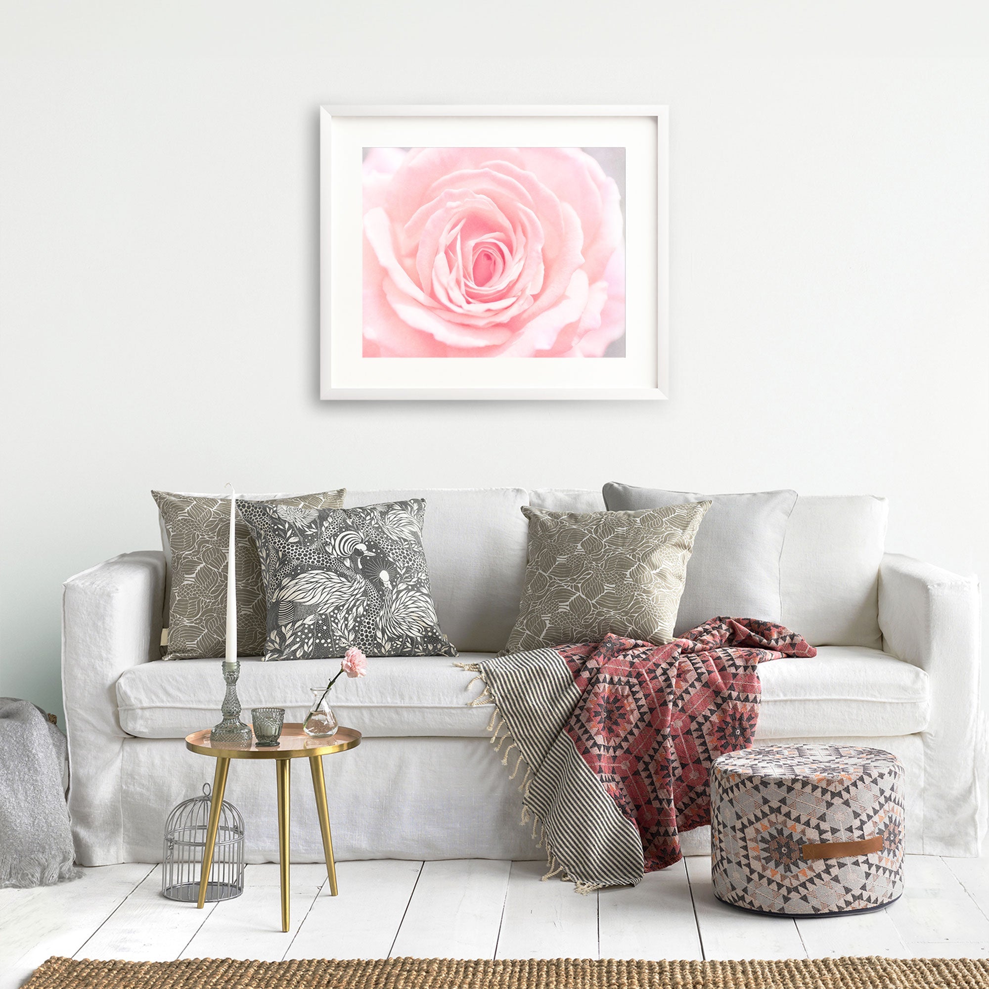 A cozy living room with a white sofa adorned with &#39;Pink Rose Print&#39; cushions from Offley Green, a framed rose in bloom artwork above, a small gold side table, and a stylish ottoman. A red patterned throw is