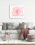 A cozy living room with a white sofa adorned with 'Pink Rose Print' cushions from Offley Green, a framed rose in bloom artwork above, a small gold side table, and a stylish ottoman. A red patterned throw is