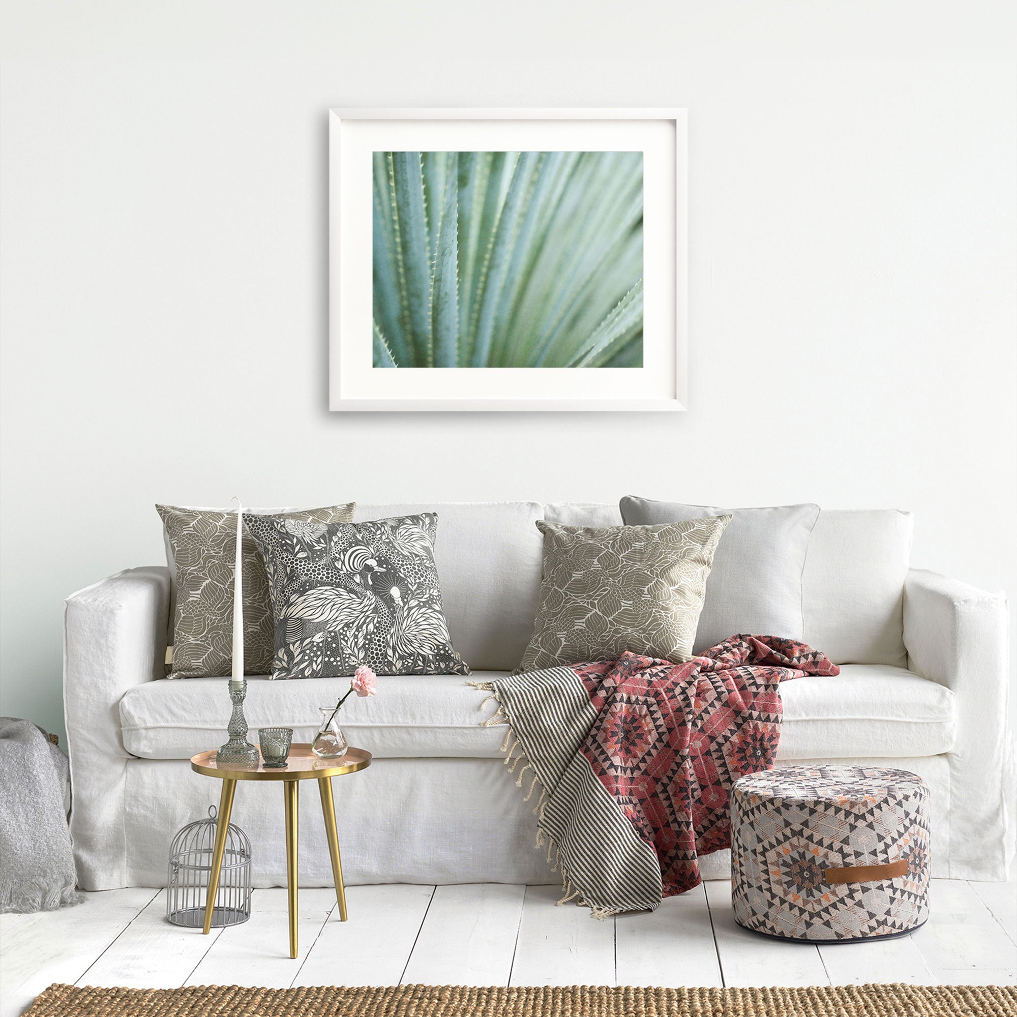 A cozy living room with a white sofa adorned with decorative pillows, a red patterned throw, a small round gold table with a vase and books, a pouf, and Offley Green's 'Strands and Spikes' Abstract Green Botanical Print.