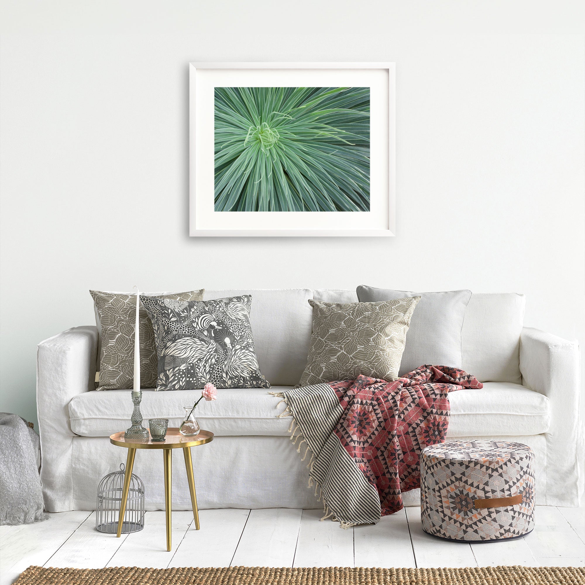 A cozy living room with a white sofa adorned with patterned throw pillows, a knitted blanket, and Abstract Green Botanical Print, 'Desert Fireworks' prints above. A small round table and decorative items complete the serene décor. (Brand Name: Offley Green)