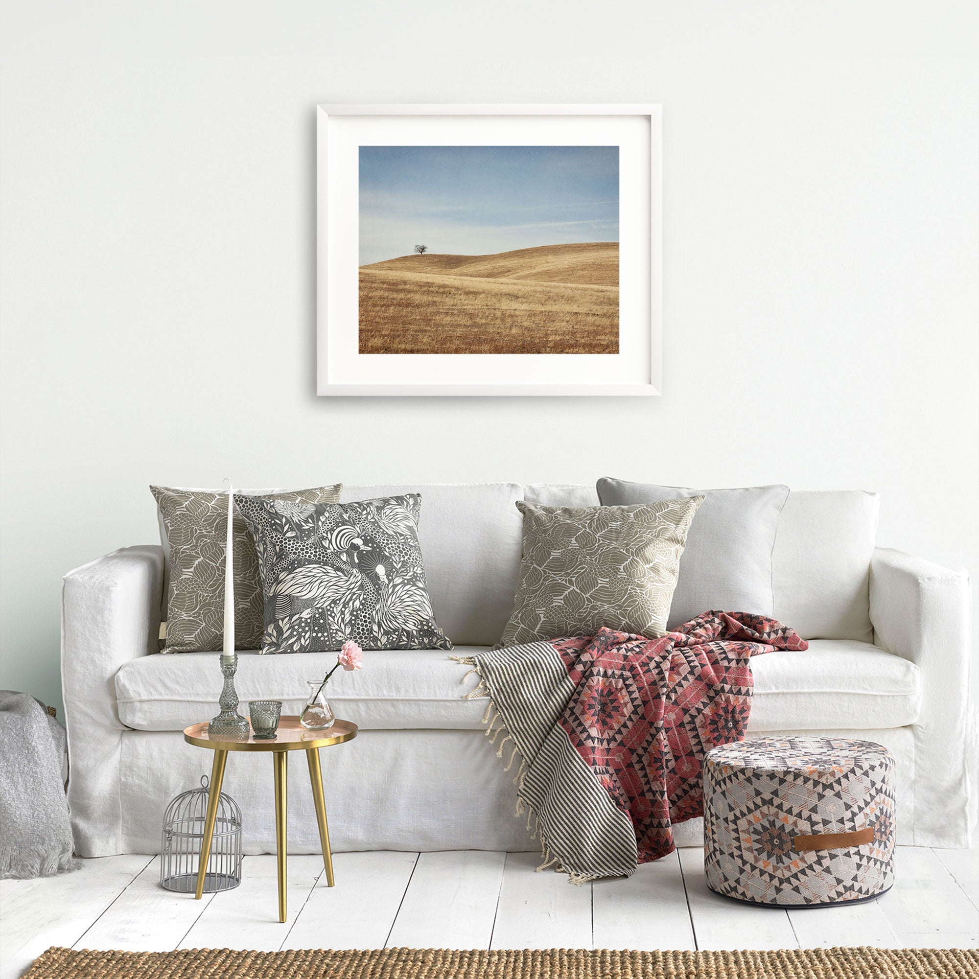 A cozy living room setup featuring a white sofa with decorative pillows, a glass side table, Offley Green California Central Coast Landscape Print 'Golden Ynez' on the wall, a red patterned throw blanket, and