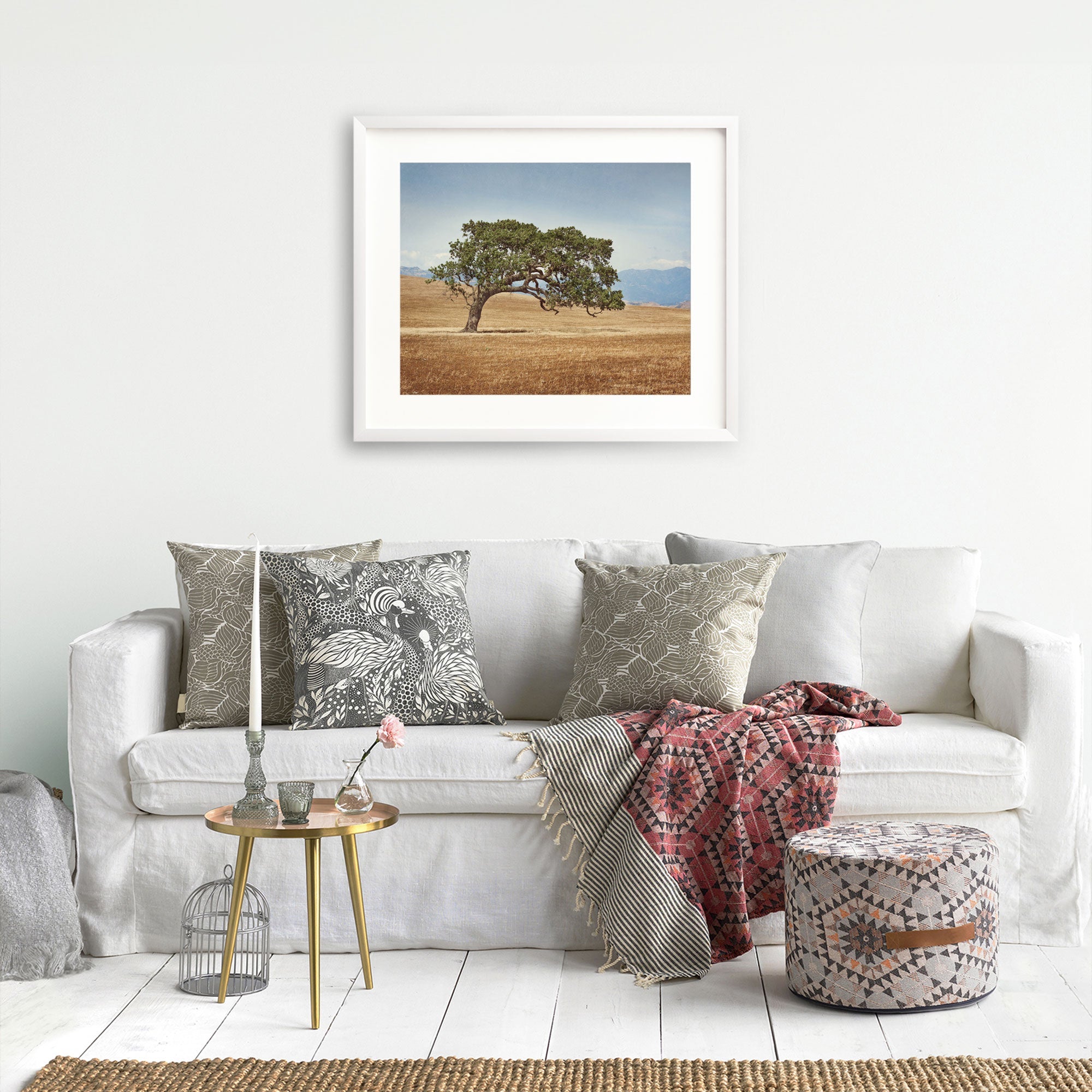 A cozy living room featuring a white sofa adorned with patterned cushions, a red throw blanket, a small round gold table with decorative items, and a framed California Oak Tree Print, 'Windswept' from Offley Green on the wall above.