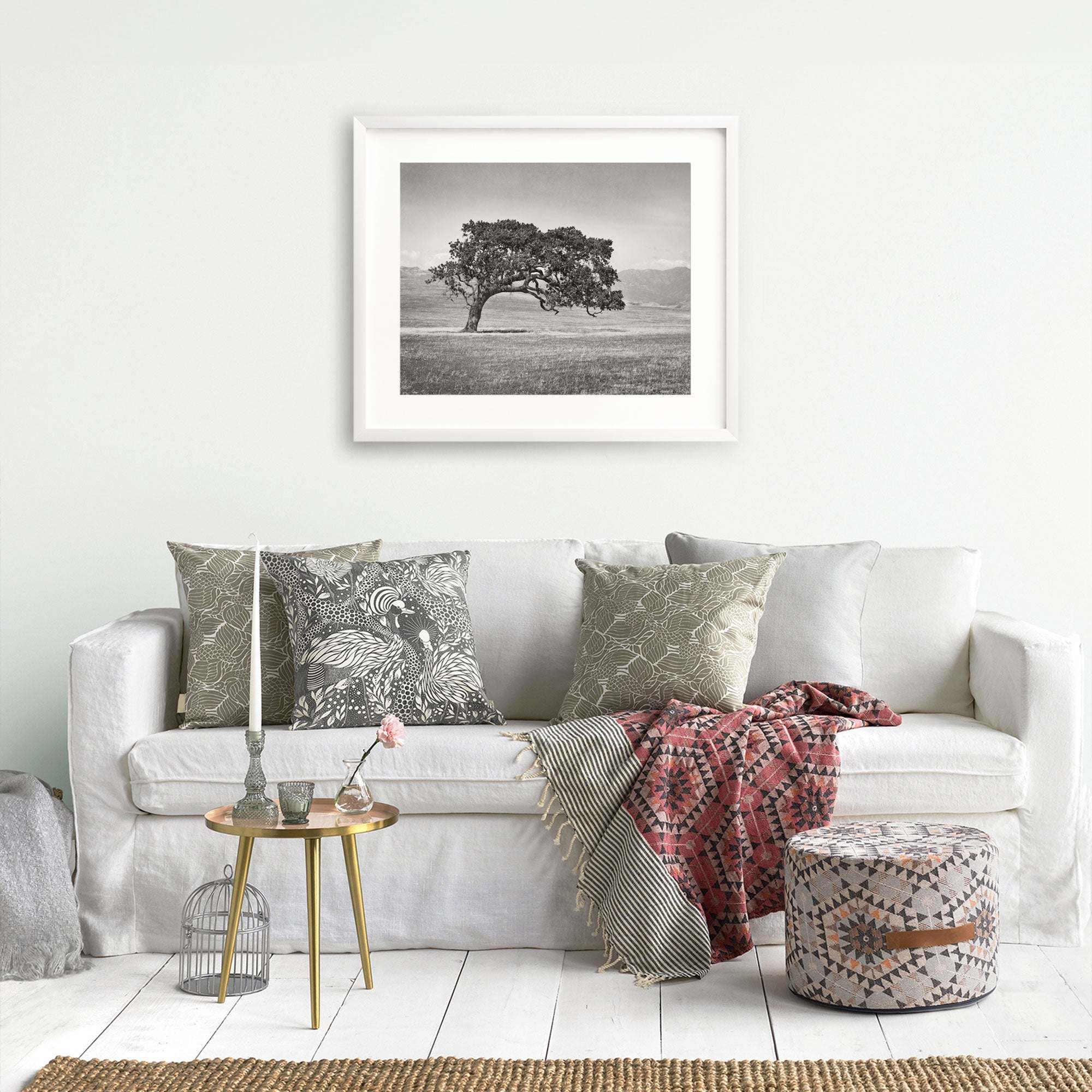 A cozy living room featuring a white sofa adorned with patterned pillows, a framed Offley Green Californian Oak Tree Landscape, 'Windswept (Black and White)' above it, a small round table with a vase, a birdcage.