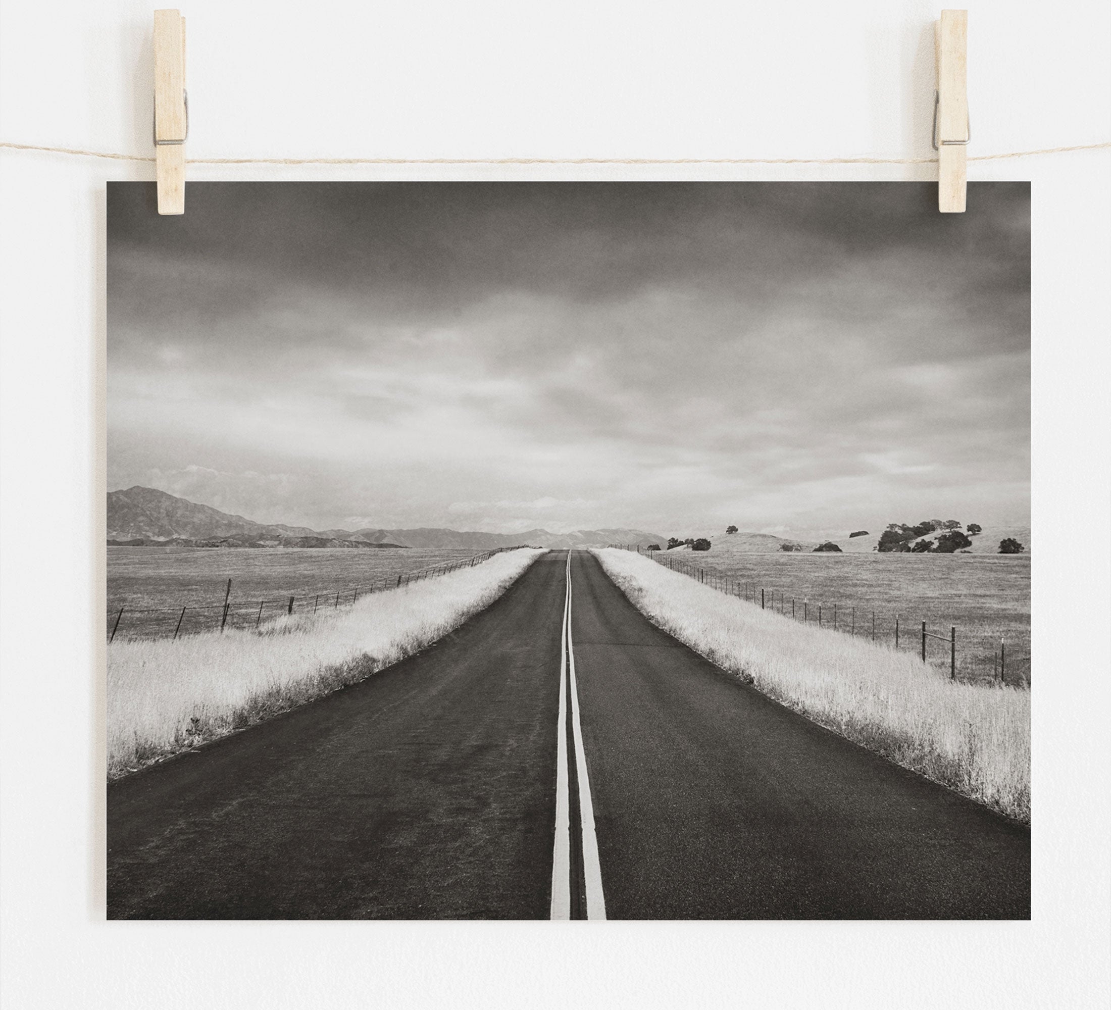 Offley Green's Black and White Rural Landscape Art, 'American Road Trip', printed on archival photographic paper, features an empty road extending into the distance under a cloudy sky, flanked by fields and mountains in sepia tones.