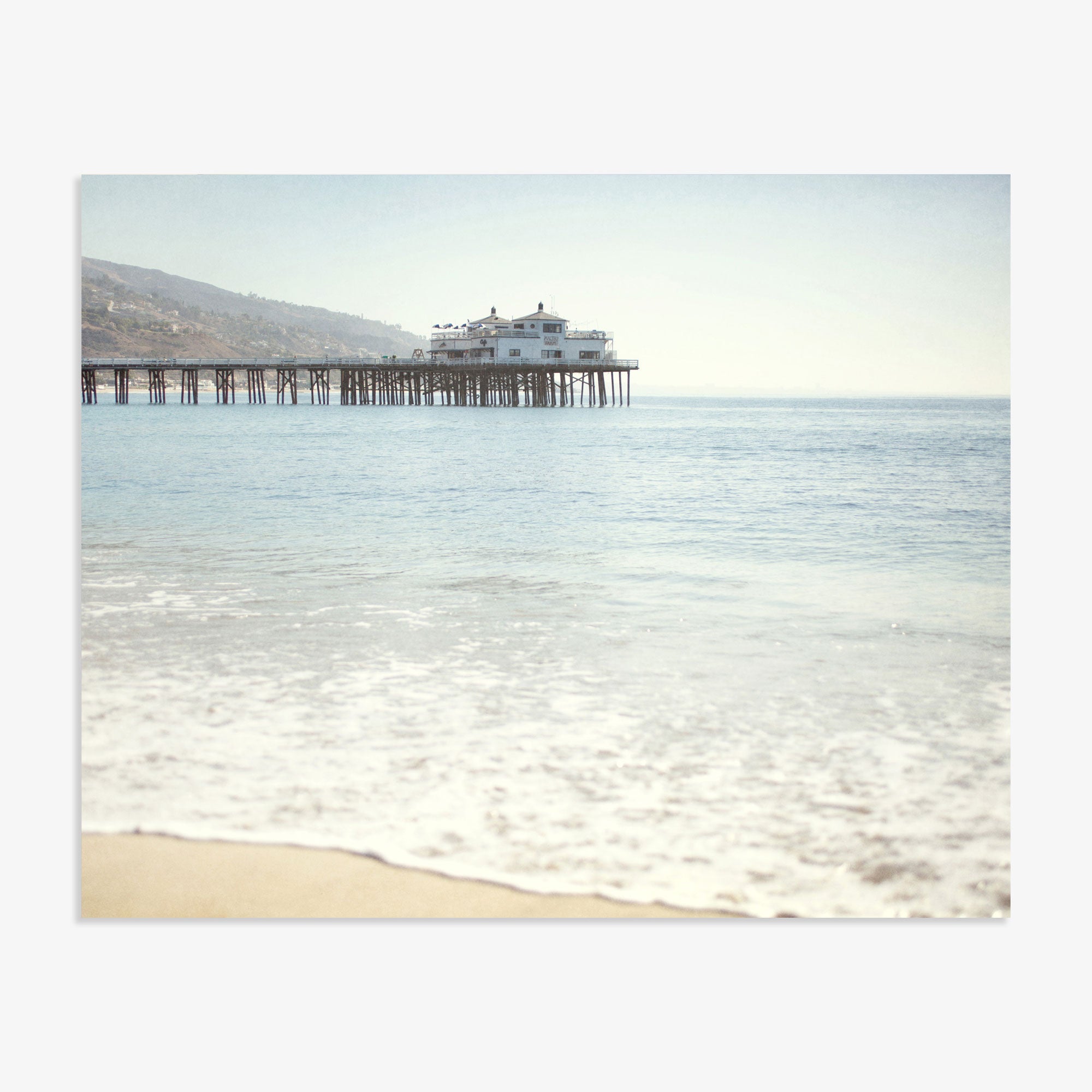 A tranquil coastal scene featuring Offley Green&#39;s &#39;Malibu Pier&#39; California Beach Print extending into the ocean, with a large building at its end under a clear blue sky. Gentle waves touch a sandy beach in the foreground.