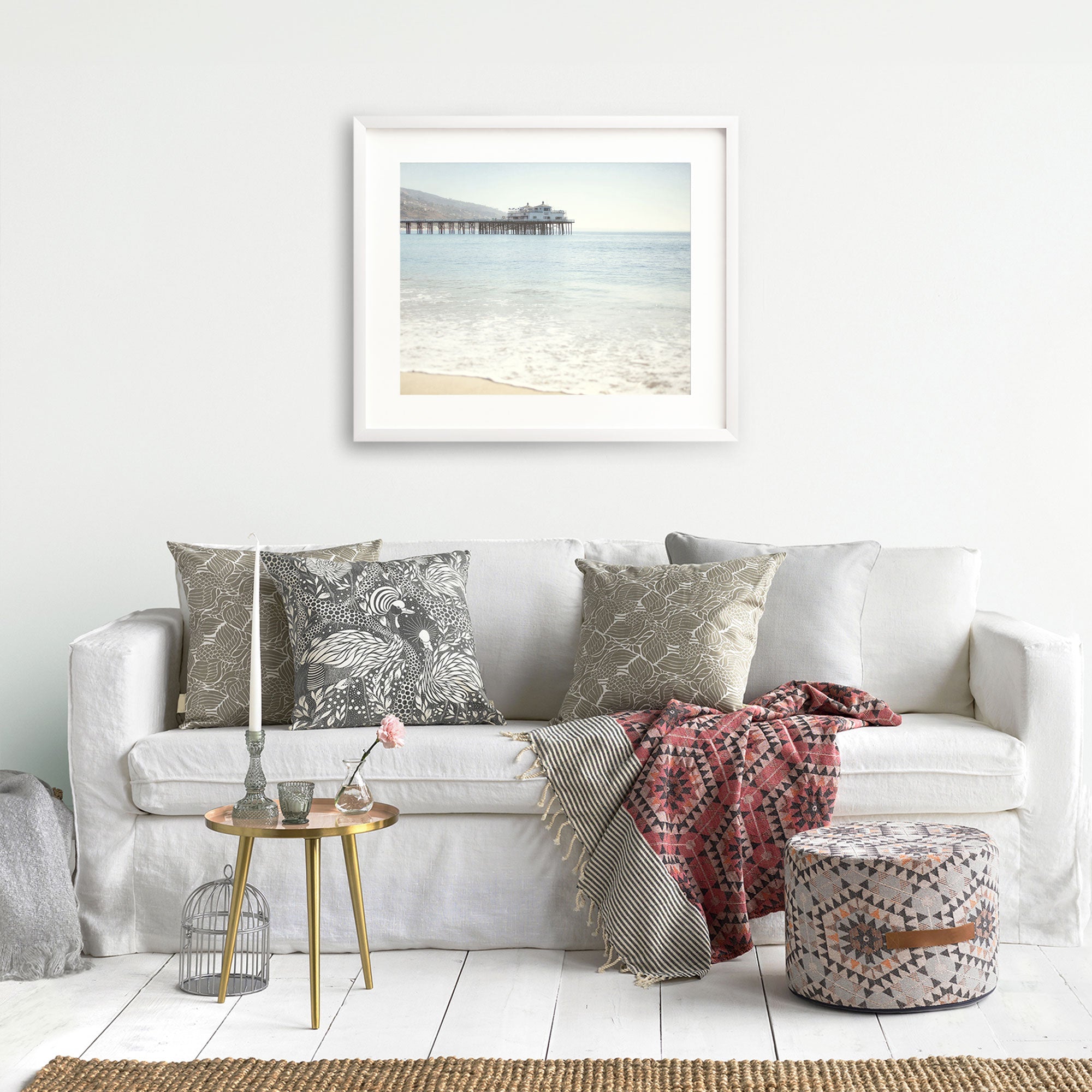 A modern living room featuring a white sofa adorned with patterned cushions, a cozy red blanket, and a small gold side table. On the wall above the sofa hangs a framed photo of California Beach Print by Offley Green.