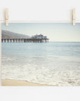 A photograph of Offley Green's 'Malibu Pier' California Beach Print extending into the ocean, pinned by wooden clothespins onto a string against a white wall. The calm sea and clear sky create a serene backdrop.
