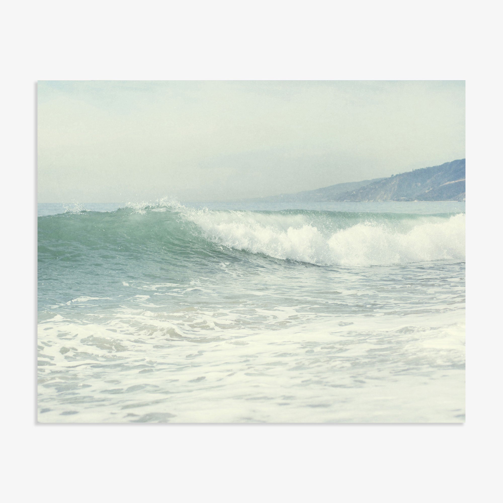 A serene Southern California beach scene depicting gentle waves rolling onto a sandy beach, with misty hills visible in the background under a soft, overcast sky. Offley Green&#39;s Coastal Print of a Breaking Wave &#39;Breaking Surf&#39;.