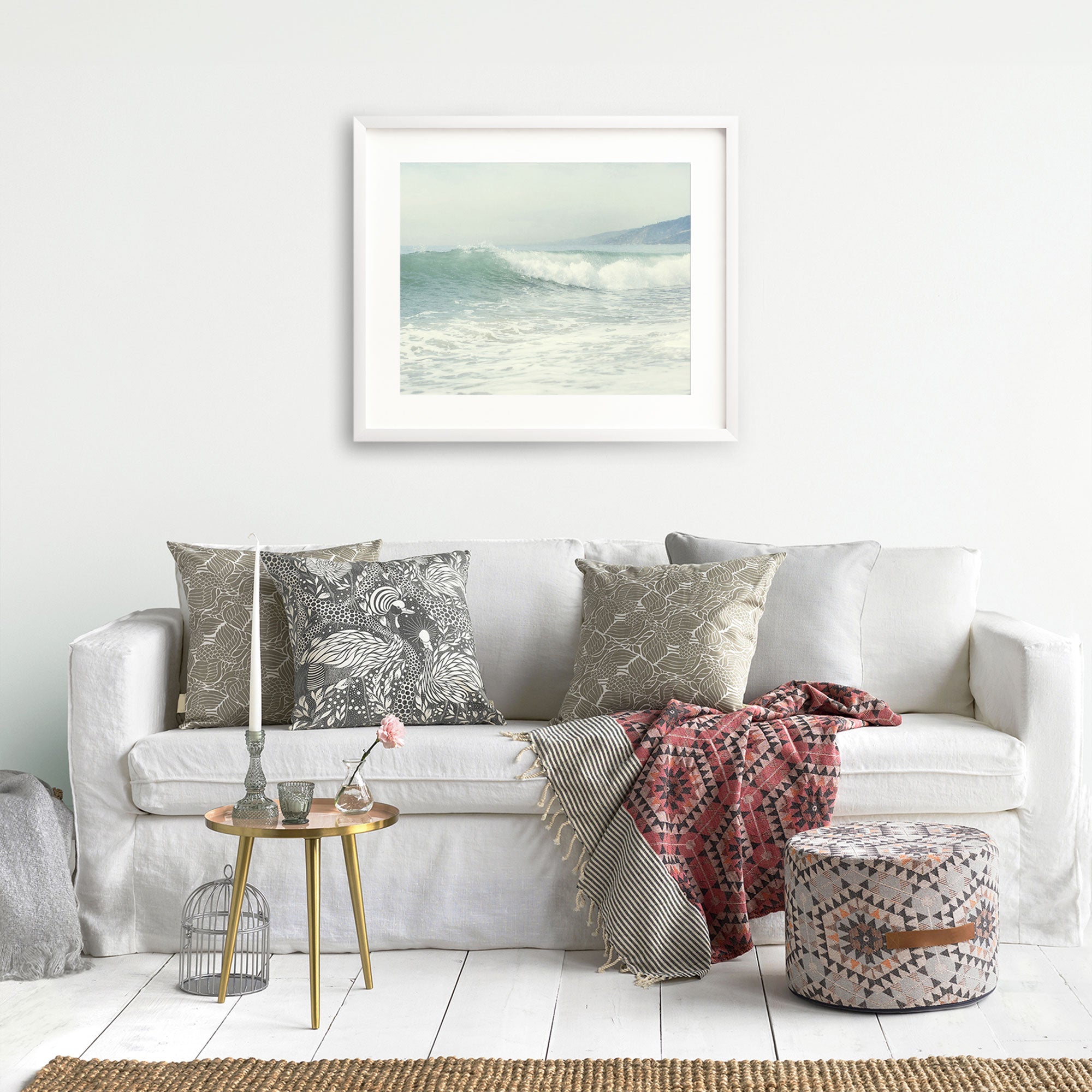 A cozy living room setup featuring a white sofa with patterned throw pillows, a draped red and gray blanket, a round gold side table with decor, a pouffe, and Offley Green's 'Breaking Surf' coastal print wall.