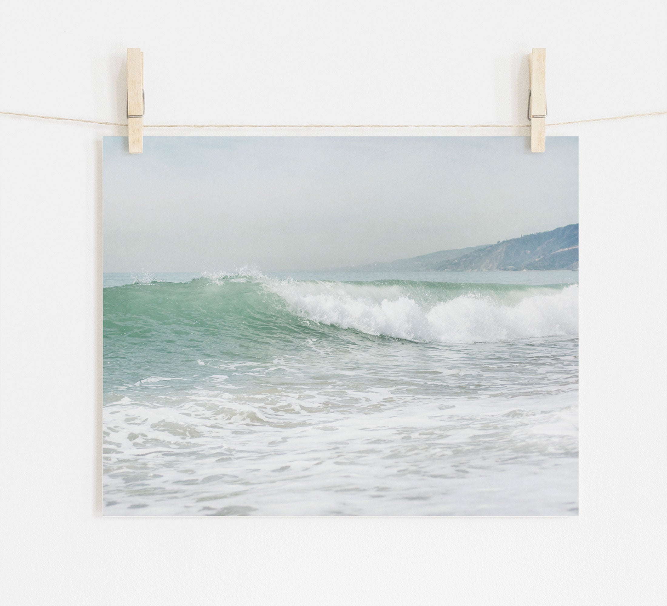 A Offley Green photo print of a breaking wave titled &#39;Breaking Surf&#39; hung on a white wall with wooden clothespins on a string, depicting a Southern California beach scene that captures gentle sea waves with a hazy coastline in the background.