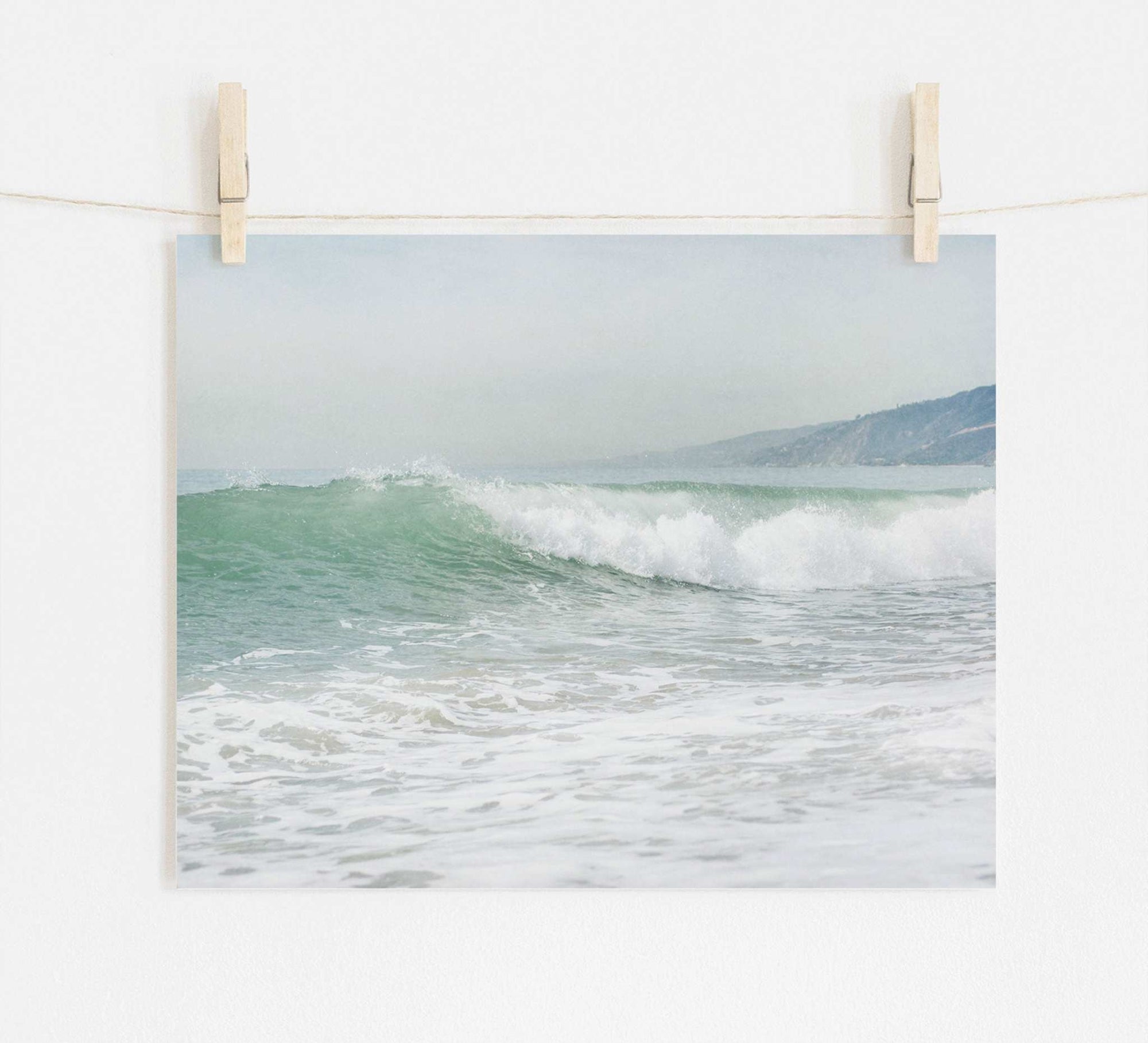 A Offley Green photo print of a breaking wave titled 'Breaking Surf' hung on a white wall with wooden clothespins on a string, depicting a Southern California beach scene that captures gentle sea waves with a hazy coastline in the background.
