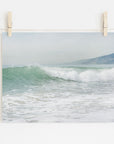 A Offley Green photo print of a breaking wave titled 'Breaking Surf' hung on a white wall with wooden clothespins on a string, depicting a Southern California beach scene that captures gentle sea waves with a hazy coastline in the background.
