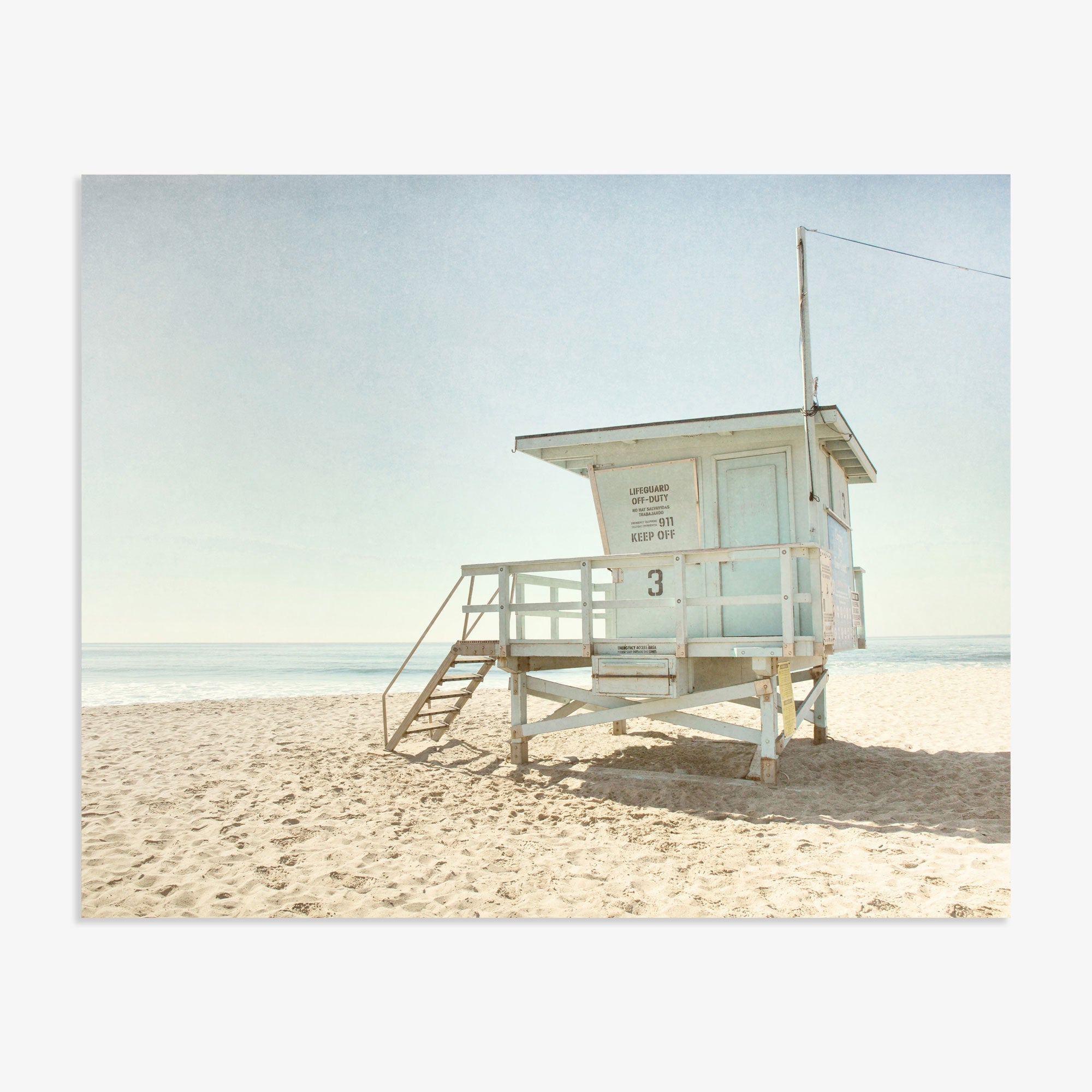 White Offley Green lifeguard tower on a sandy Malibu beach under a clear sky, with no people visible. The tower features steps leading up to an enclosed area.