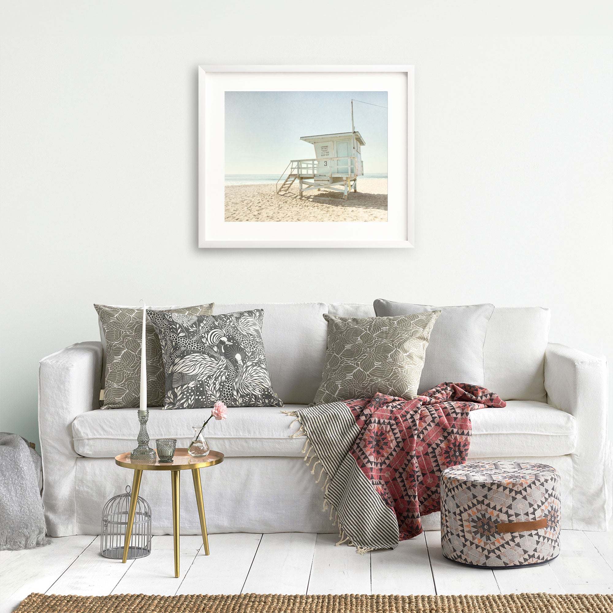 A cozy living room setting featuring a white sofa adorned with patterned pillows, a decorative throw, a small side table with a flower vase, and an unframed beach photograph of lifeguard towers in California Summer Beach Art by Offley Green.