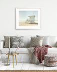 A cozy living room setting featuring a white sofa adorned with patterned pillows, a decorative throw, a small side table with a flower vase, and an unframed beach photograph of lifeguard towers in California Summer Beach Art by Offley Green.