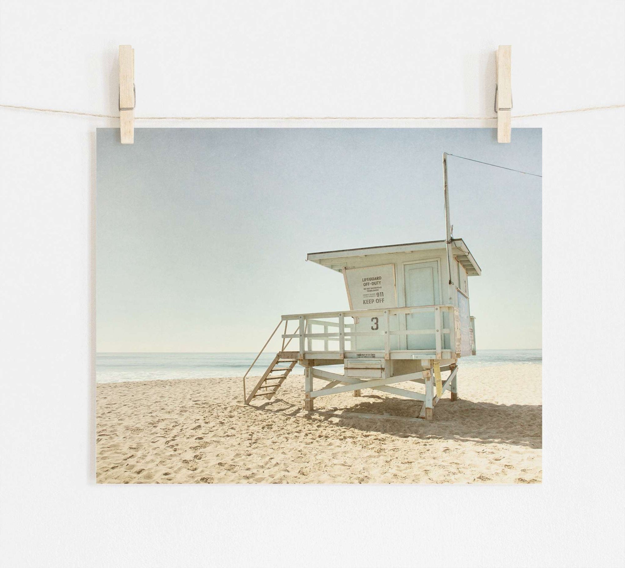 A photo of Offley Green's California Summer Beach Art, 'Malibu Lifeguard Tower', numbered "3" on Malibu beach, pinned by wooden clothespins on a line, set against a scene with soft sand and a clear sky.